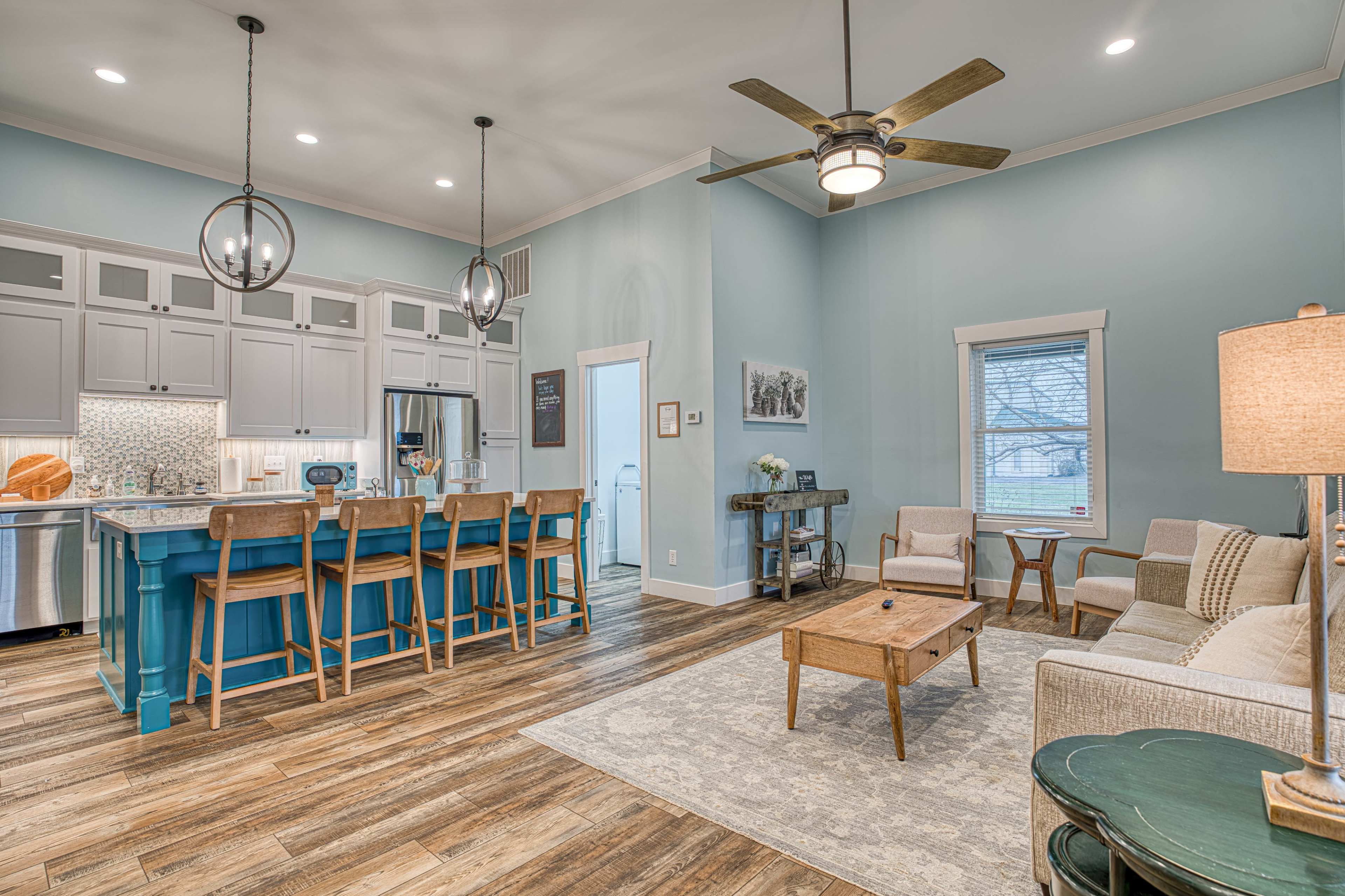 The image shows a modern living room and kitchen area with light blue walls, wooden flooring, and a kitchen island with bar stools.