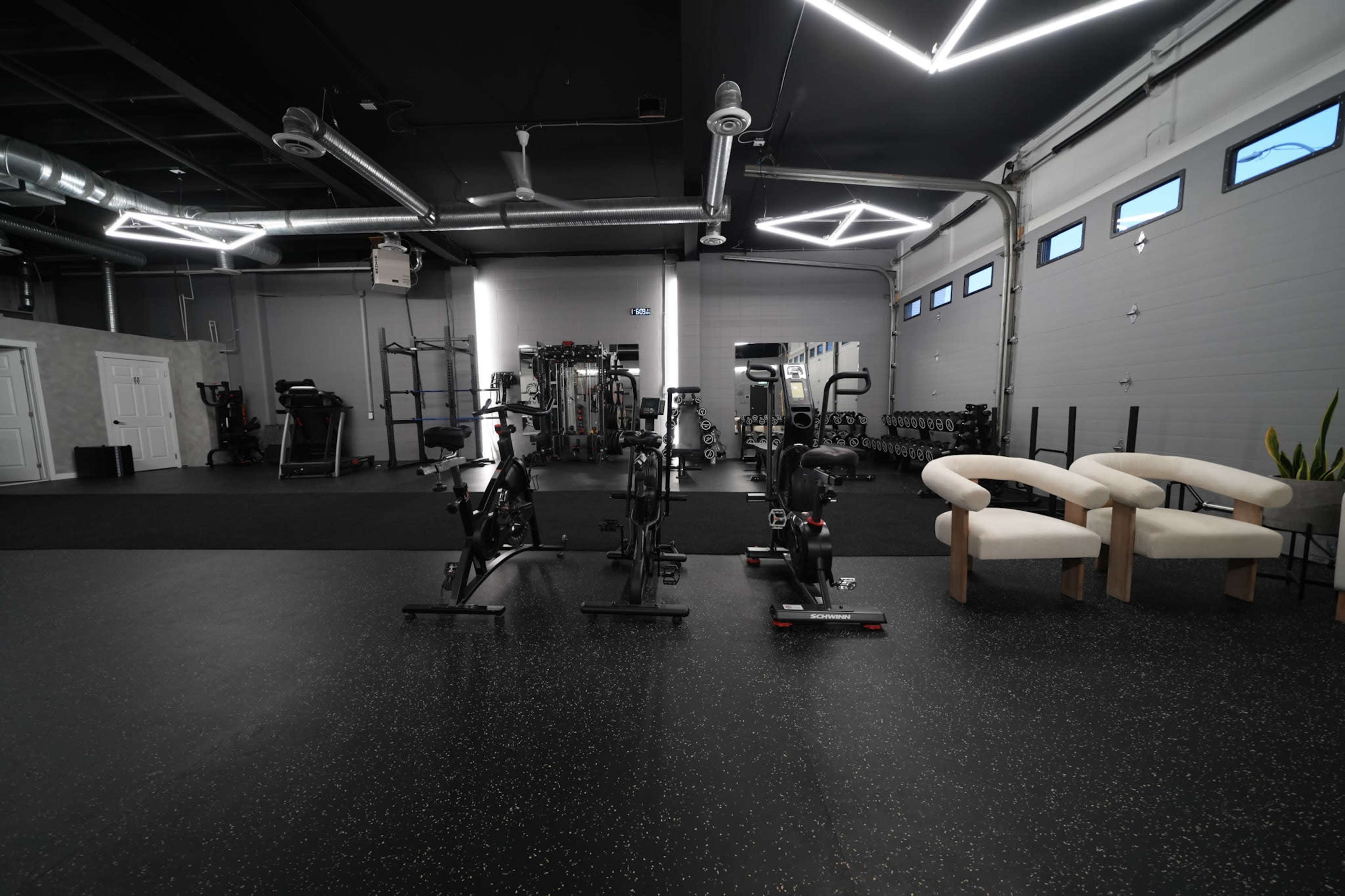 The image shows a spacious gym interior equipped with various exercise machines, free weights, and two modern chairs situated on a rubberized floor.
