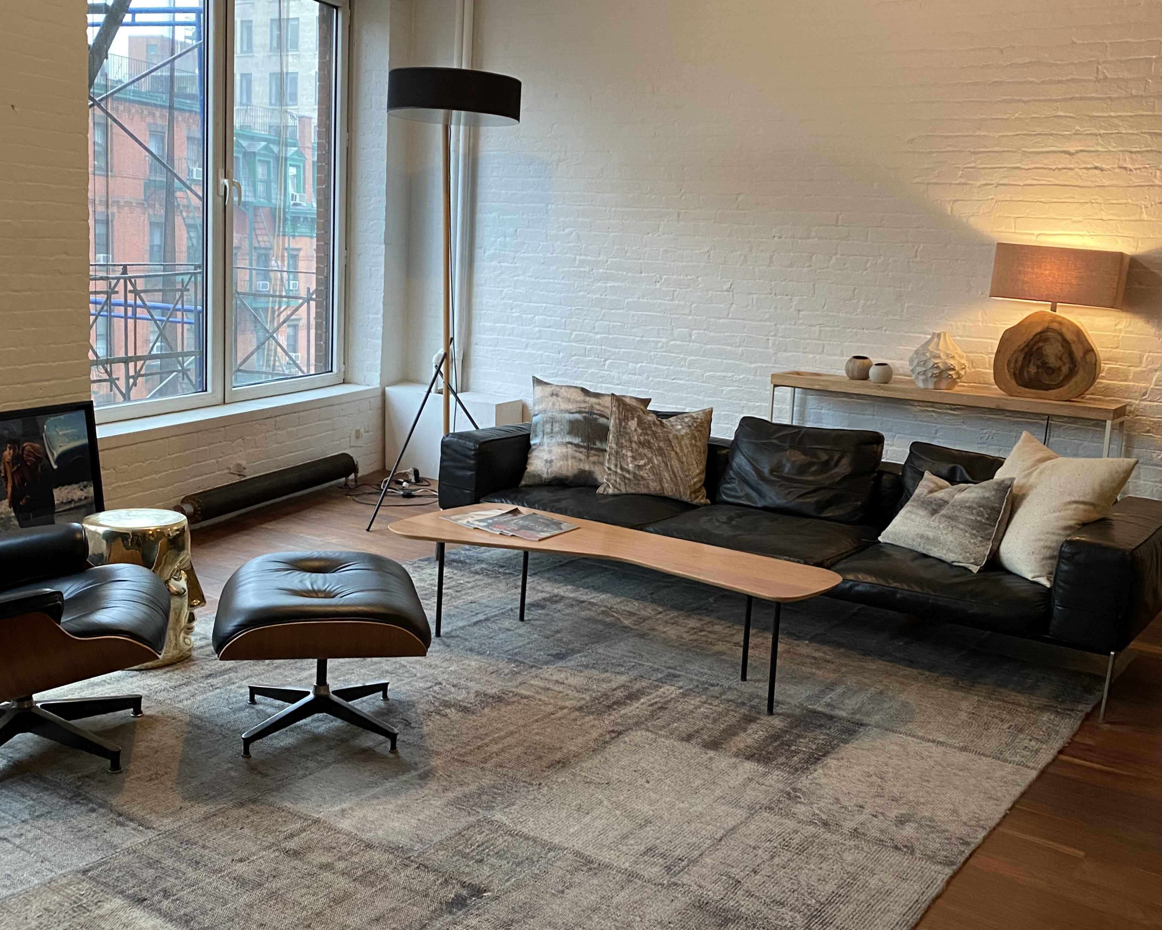 A modern living room featuring a black leather sofa with decorative pillows, a wooden coffee table, an ottoman, and a floor lamp, all set against a backdrop of large windows.