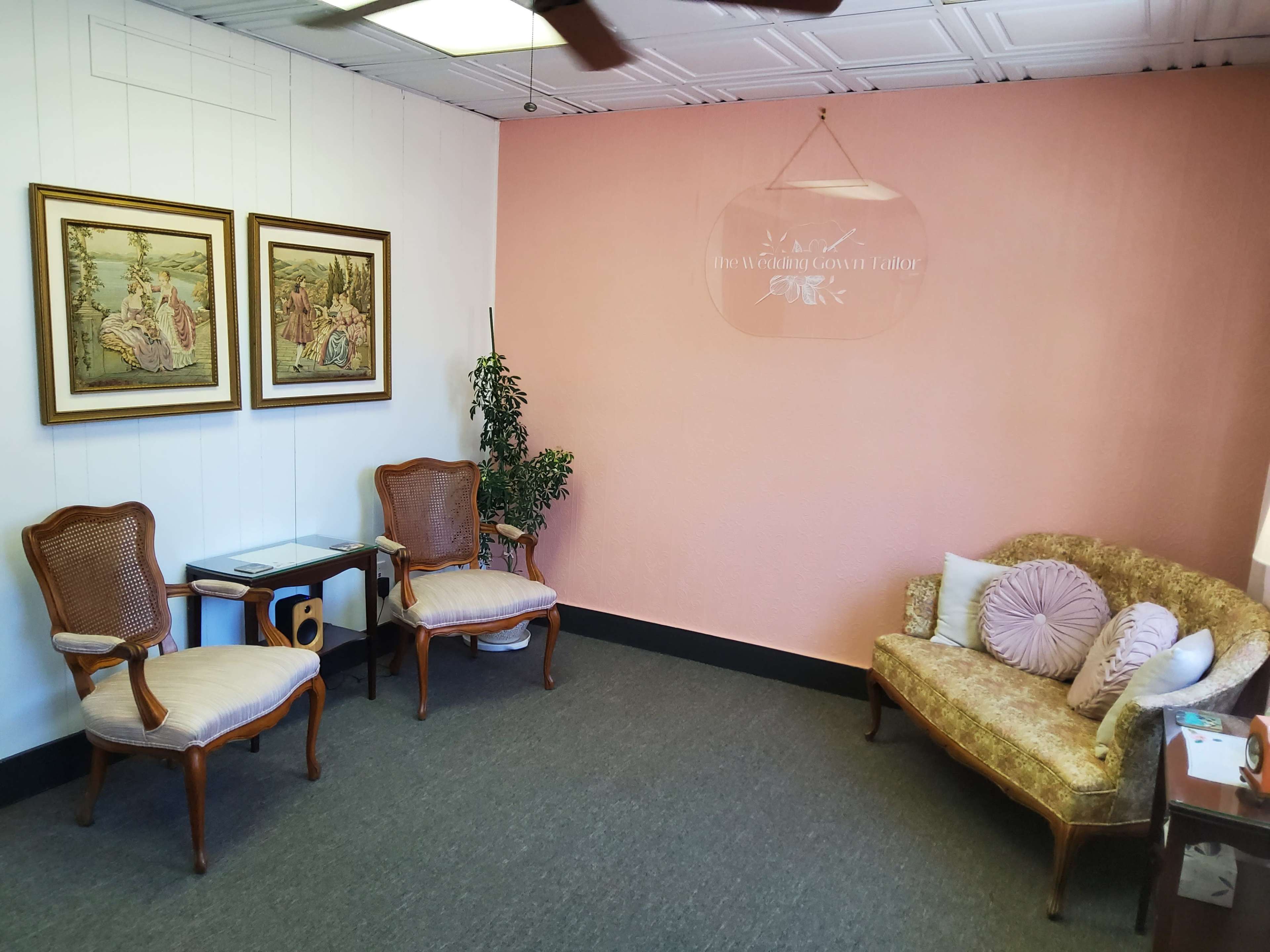 A small, softly lit room with two wooden chairs, a small table, and a couch against a pink wall, adorned with framed artwork.