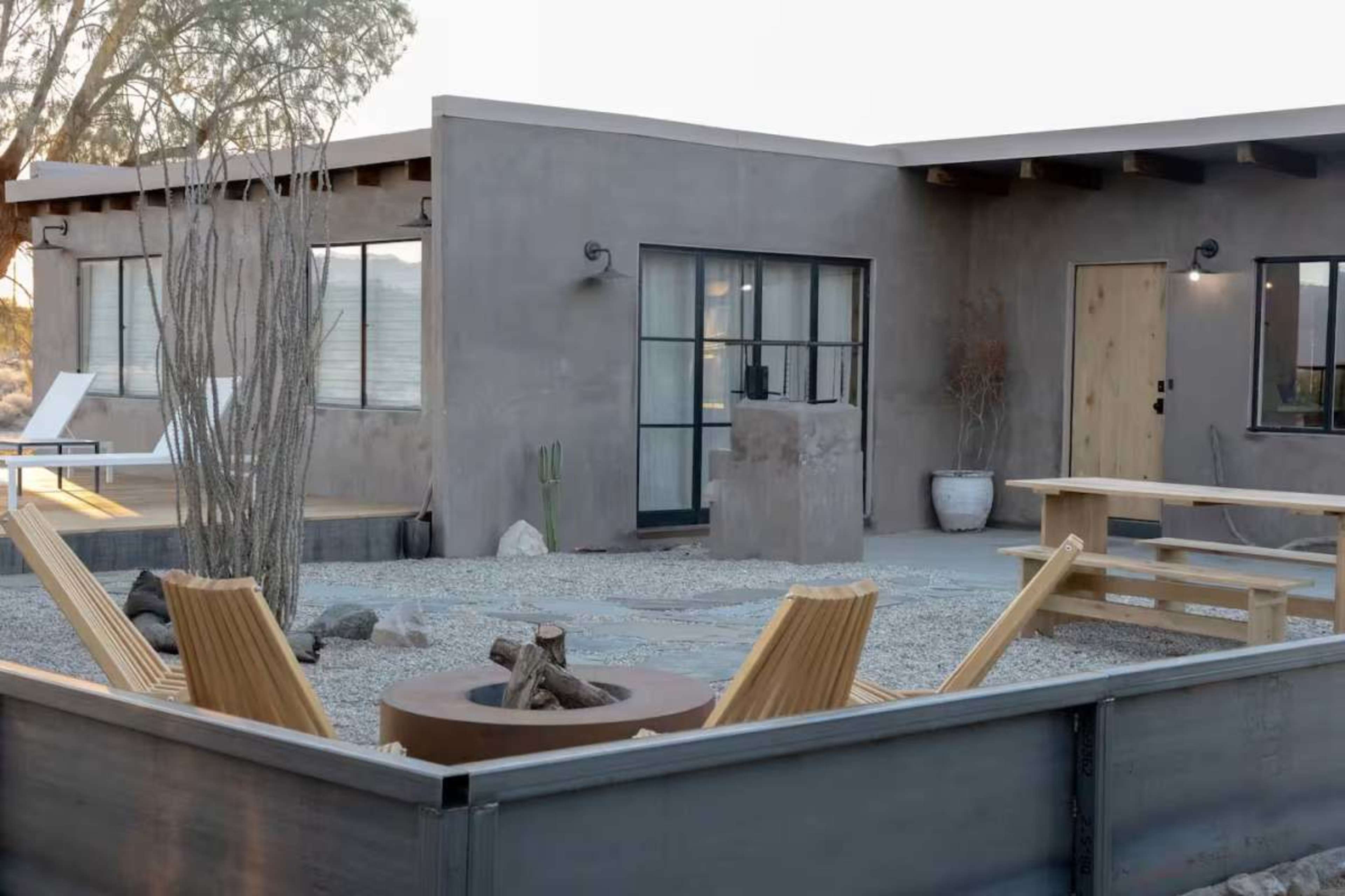 The image shows a modern adobe-style home with a landscaped outdoor area featuring seating, a fire pit, and desert vegetation.