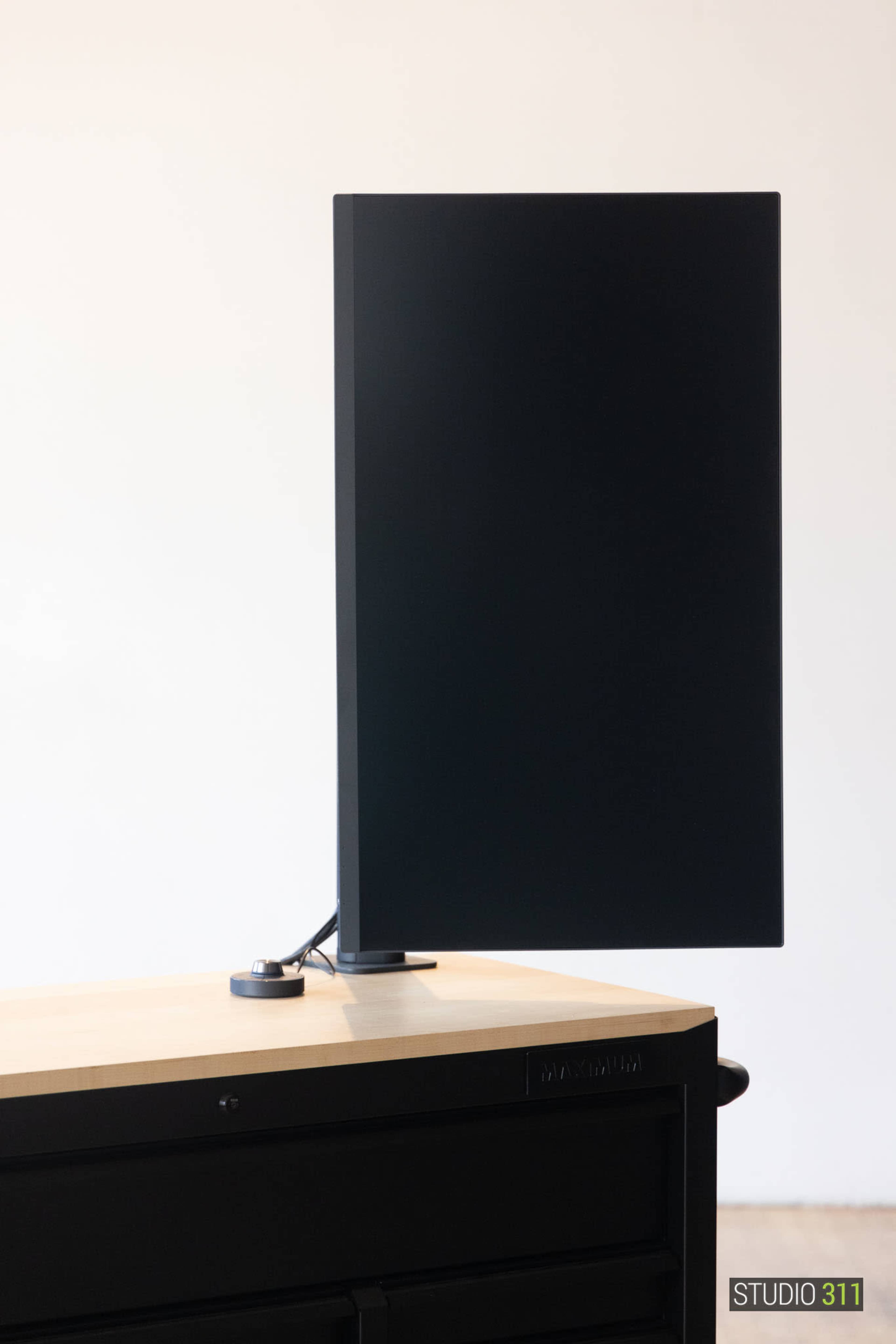 A vertical monitor is mounted on a sleek stand atop a wooden desk with a dark storage unit below.