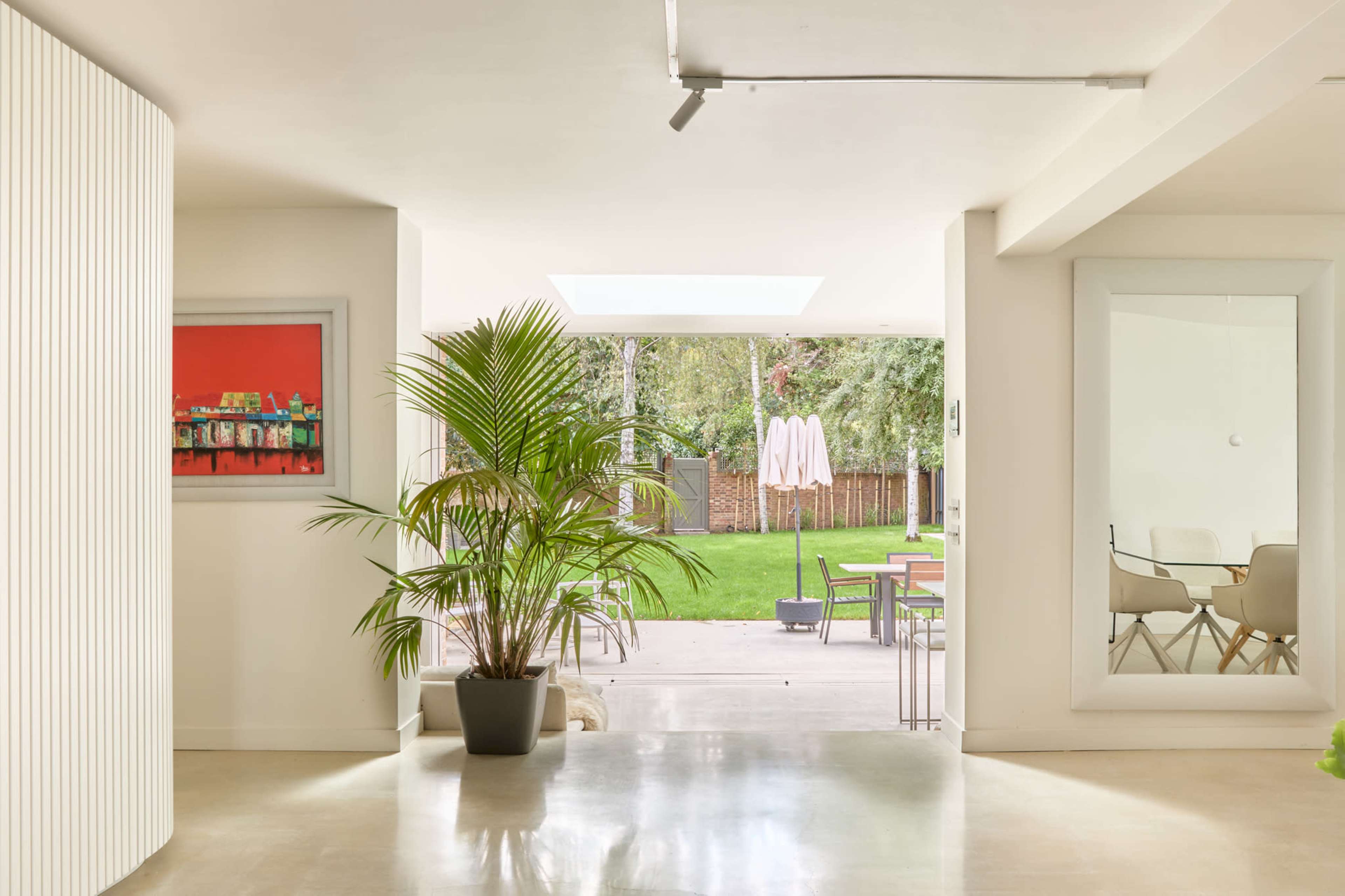 The image shows a bright, modern interior with white walls, a large window opening to a green outdoor space, and a potted plant near a contemporary artwork.