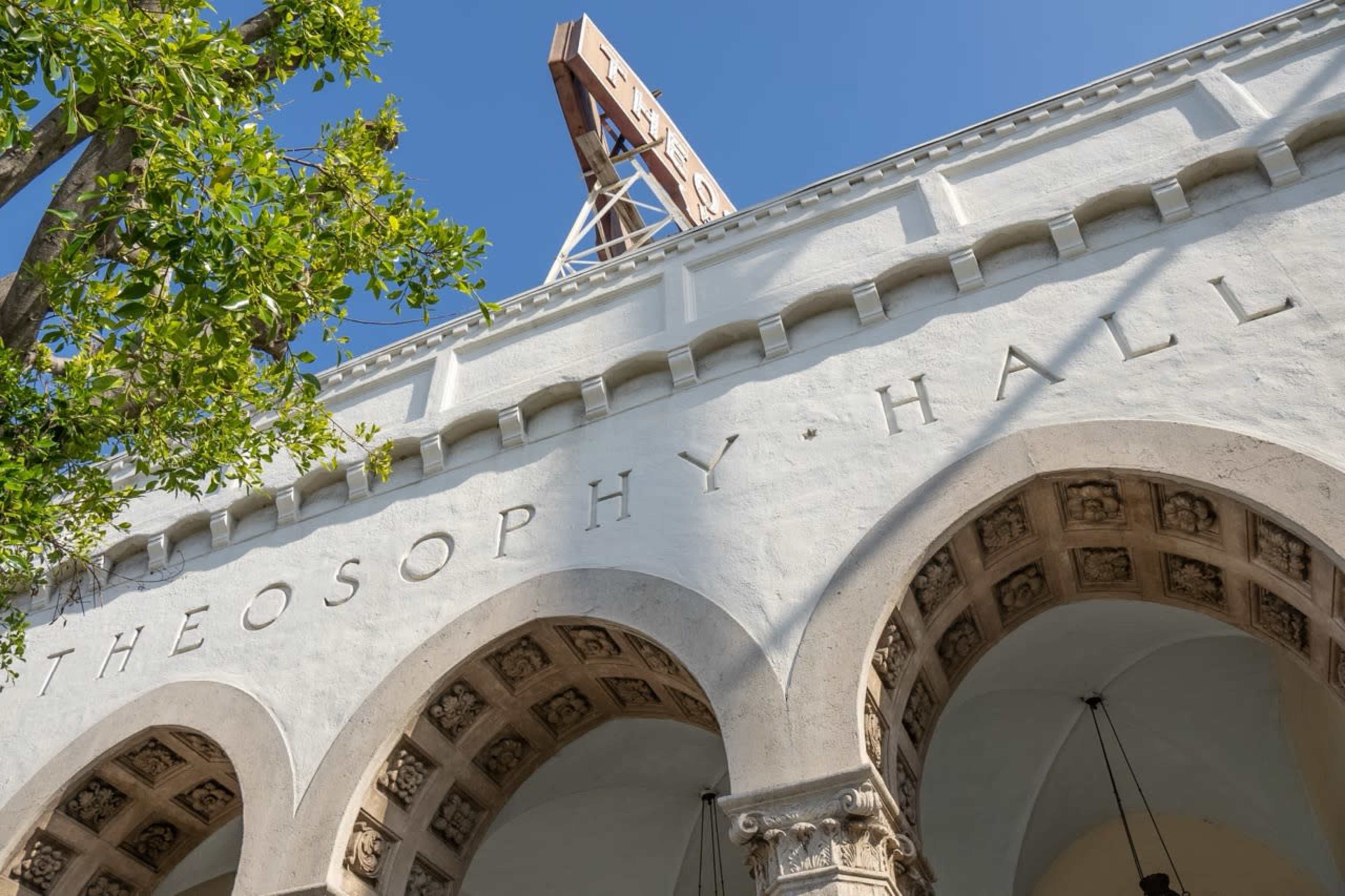 The image shows the exterior of a building with an arched design, labeled "THEOSOPHY HALL," beneath a clear blue sky and a sign atop the structure.