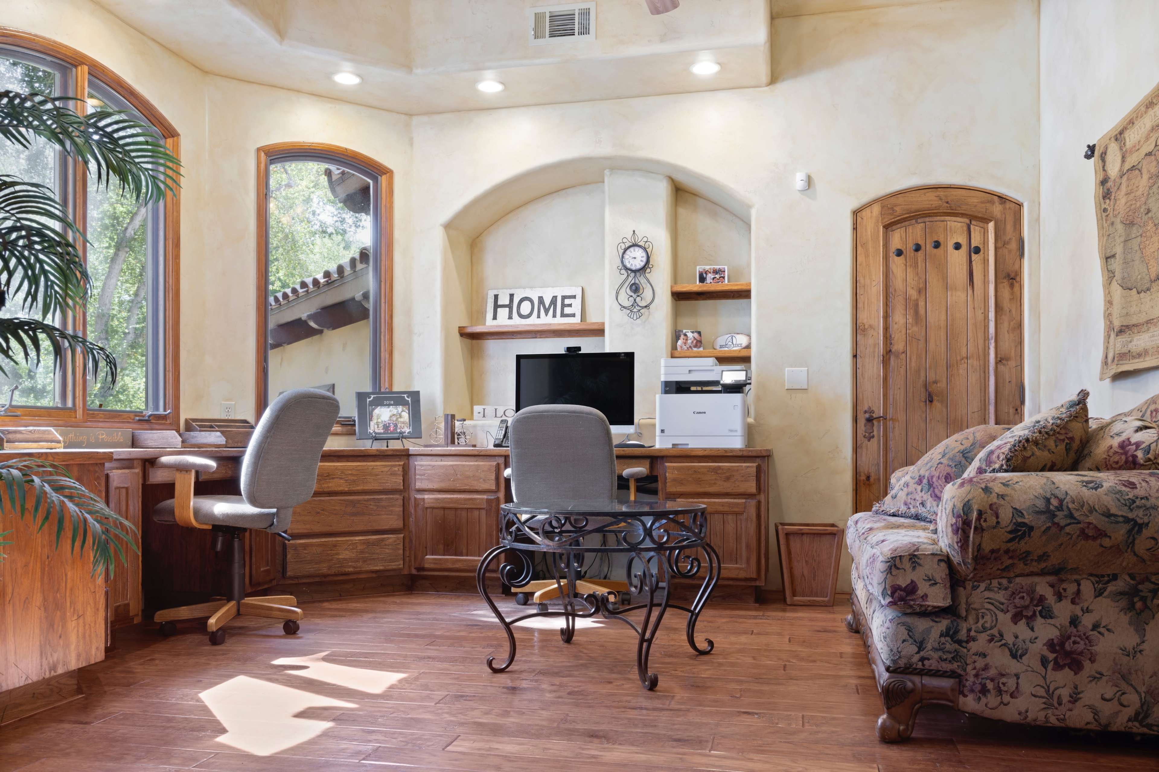 A home office features two chairs at a wooden desk with a computer, printer, and decorative elements in a well-lit room with large windows.