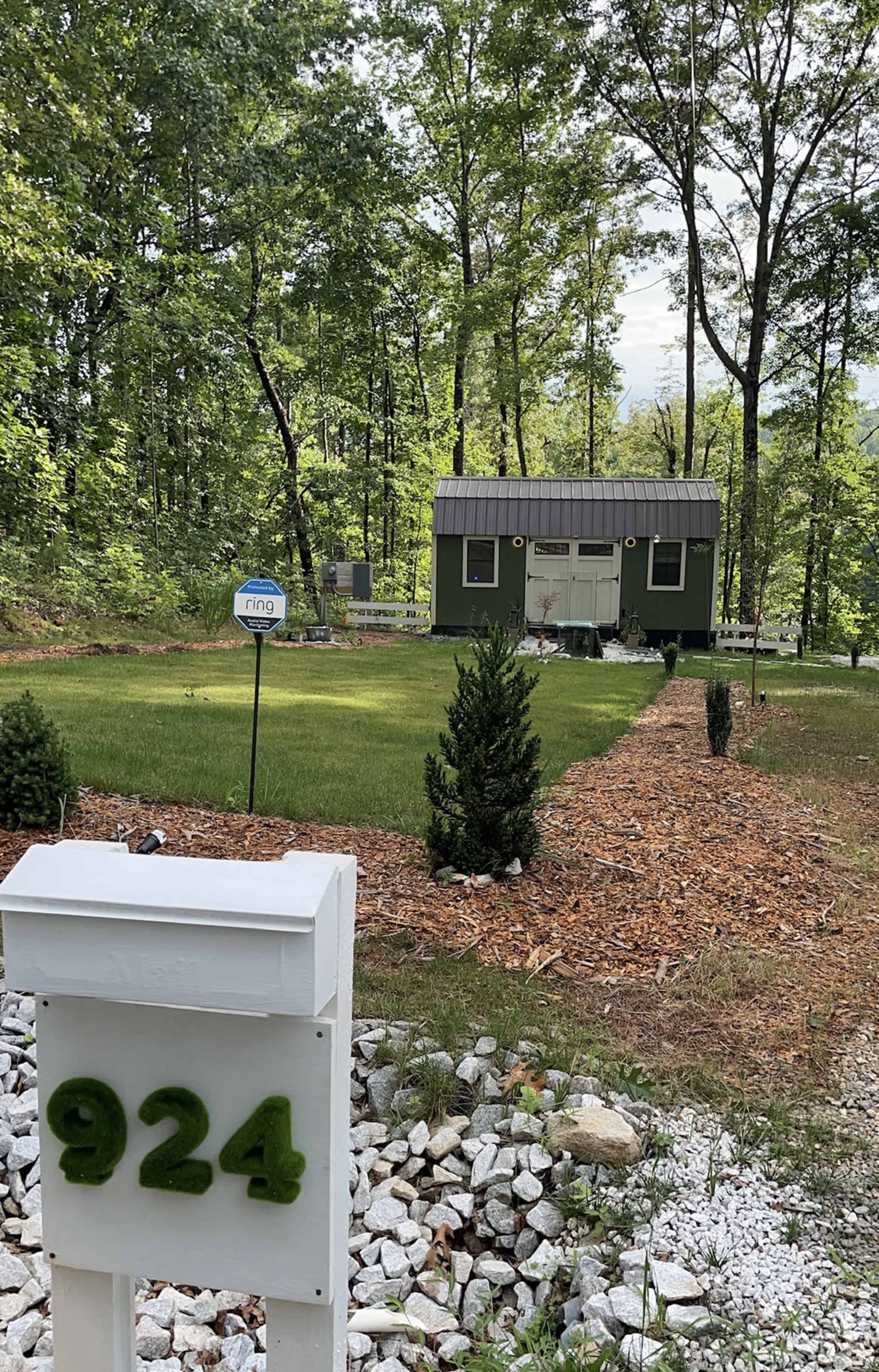 A small green cabin with a metal roof is surrounded by a grassy yard and trees, with a mailbox marked "924" in the foreground.