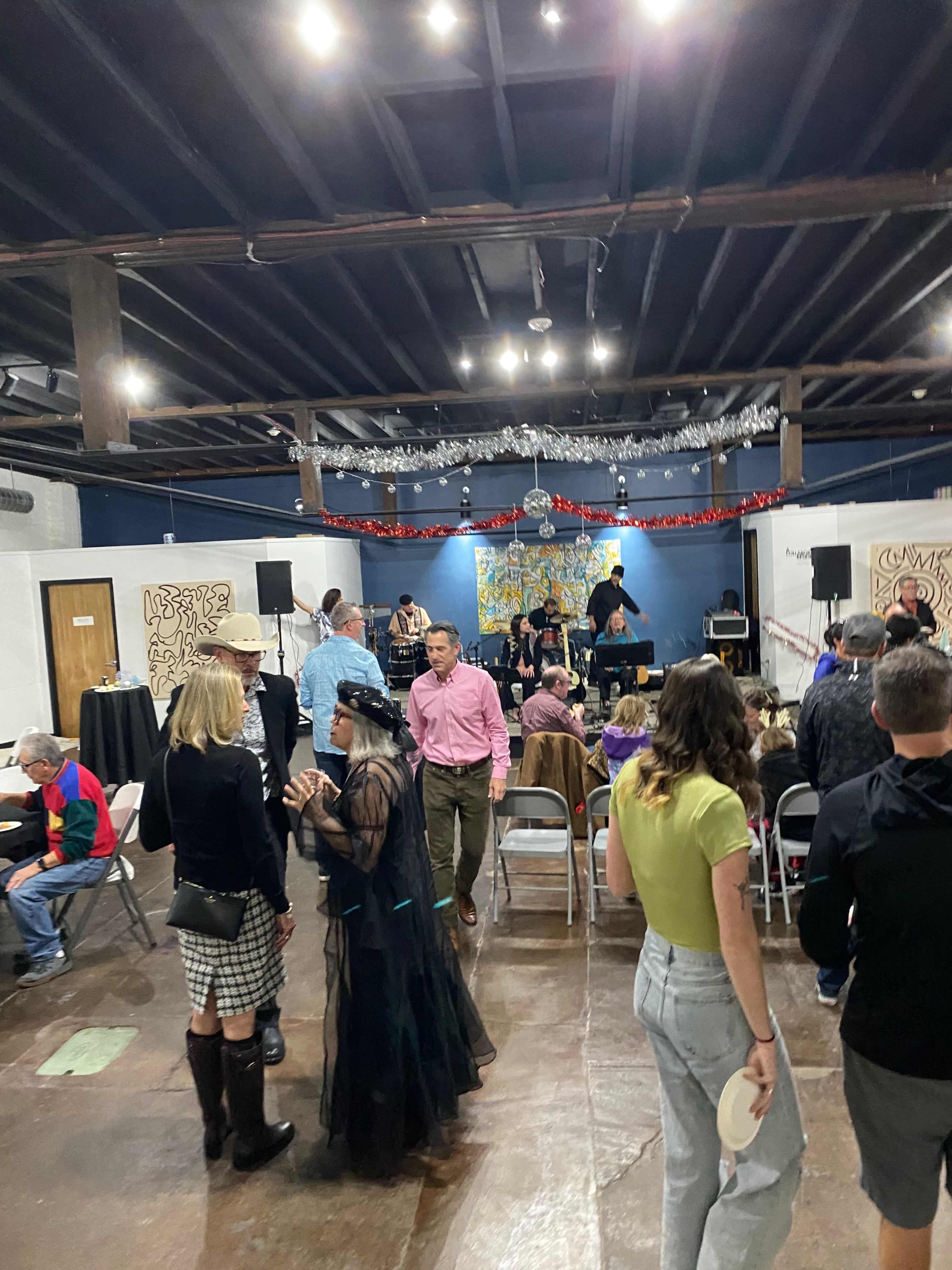 A lively gathering takes place in a spacious indoor venue, featuring people socializing, a live band performing, and colorful decorations overhead.