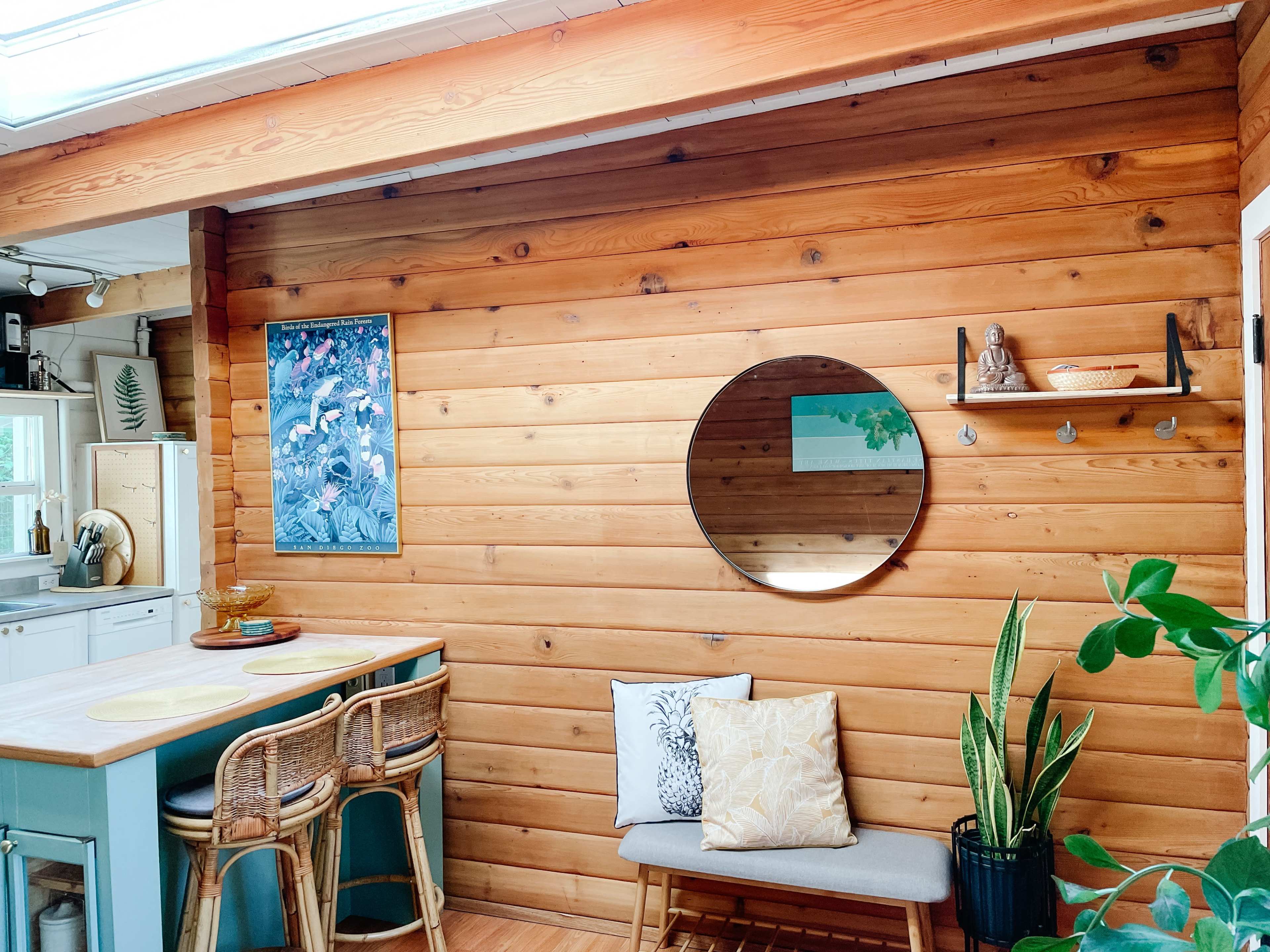 A wooden interior wall, a round mirror, a shelf with decorative items, and a small seating area with cushions beneath a large window.