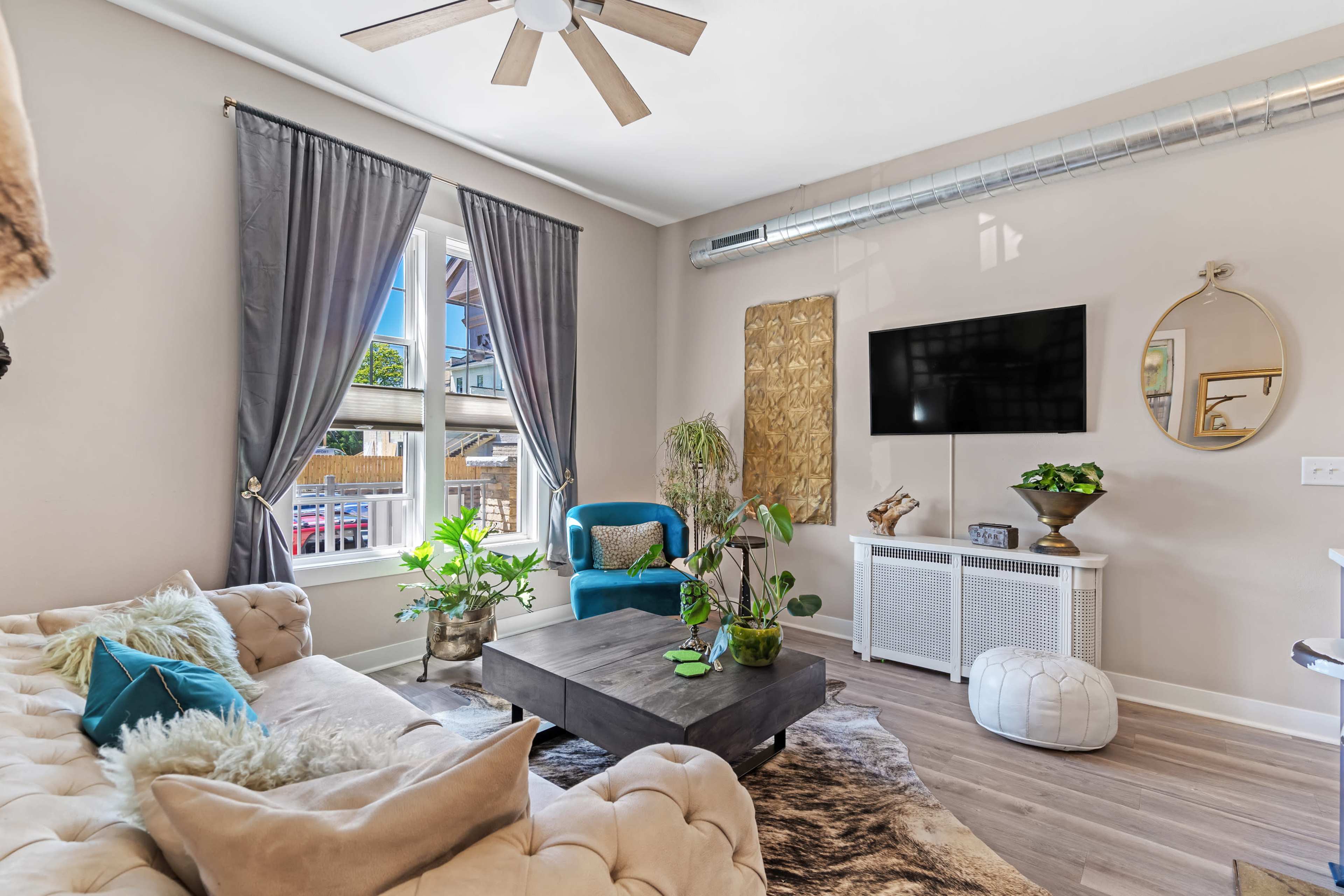 A cozy living room with a tufted sofa, a wooden coffee table, plants, and a mounted TV against a neutral backdrop.