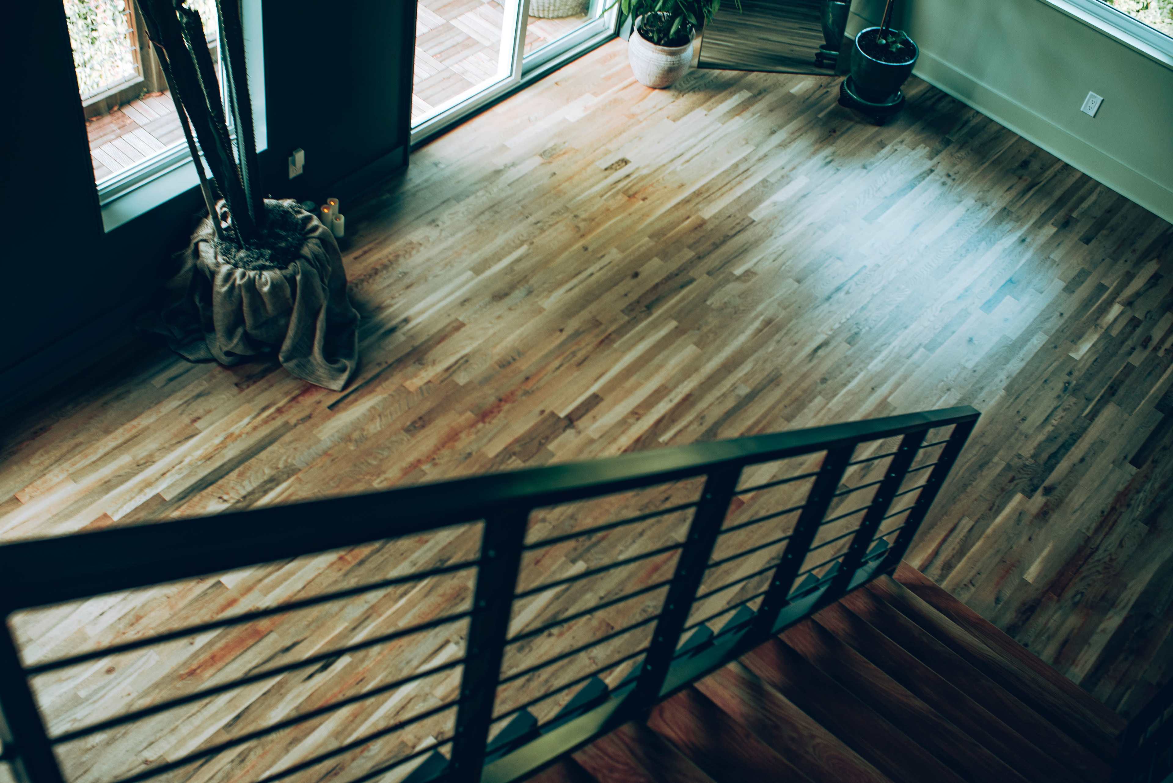 A wooden staircase leads down to a spacious room with large windows and plants in the corners.