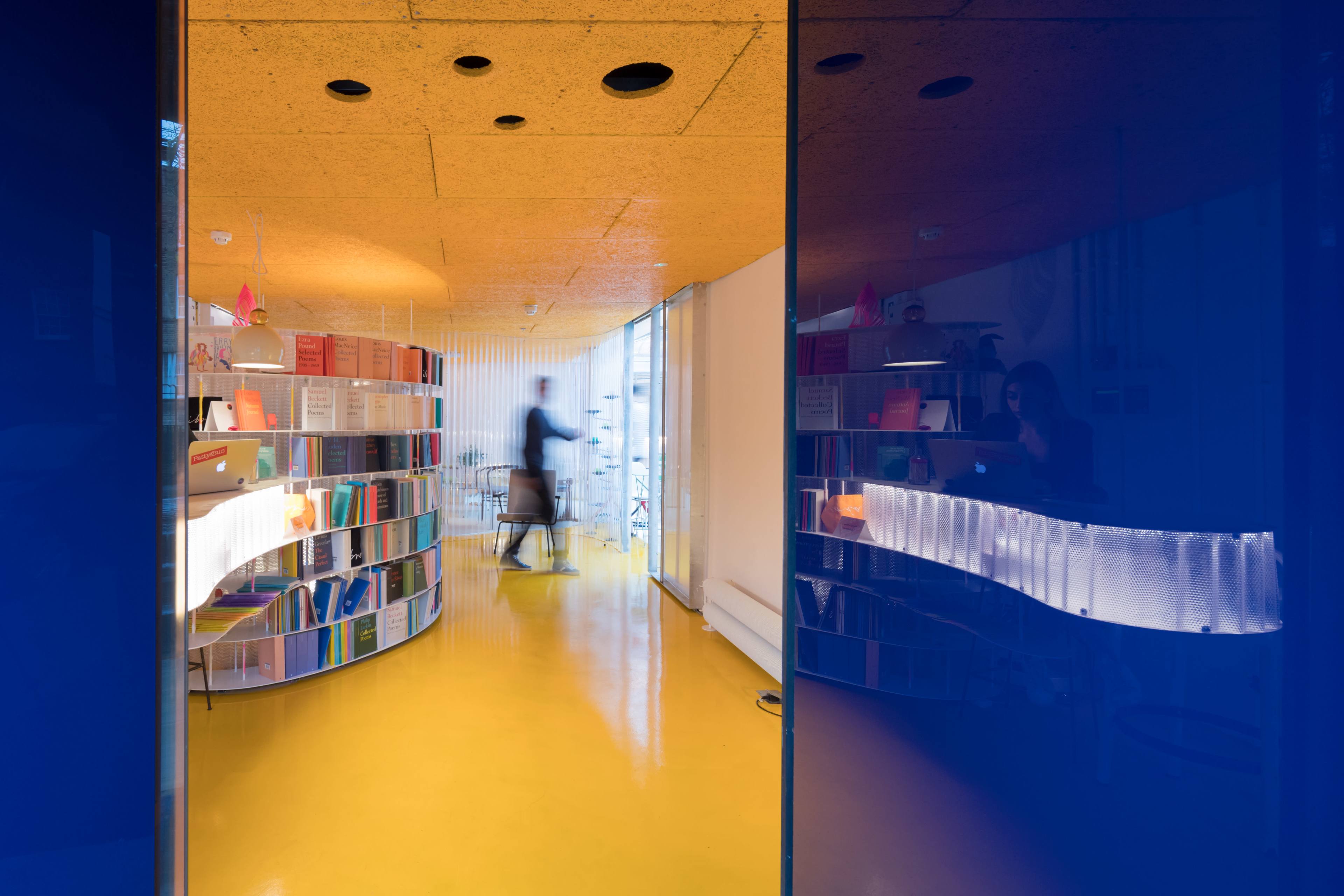 A modern office space features a circular bookshelf filled with colorful books against a backdrop of yellow flooring and a glass partition.