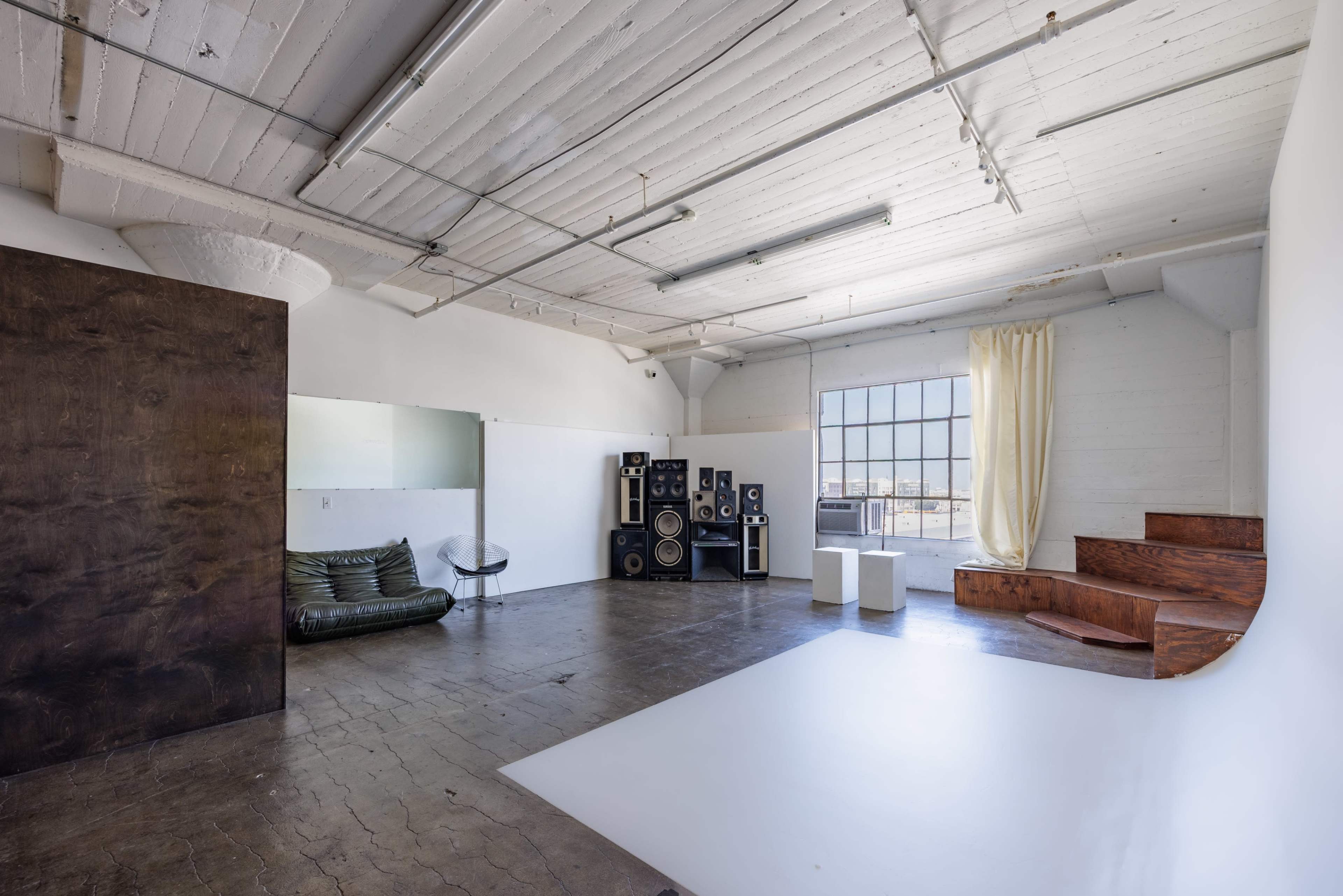 The image depicts a spacious, modern interior studio with polished concrete floors, large windows, and a mix of minimalist furniture and equipment.