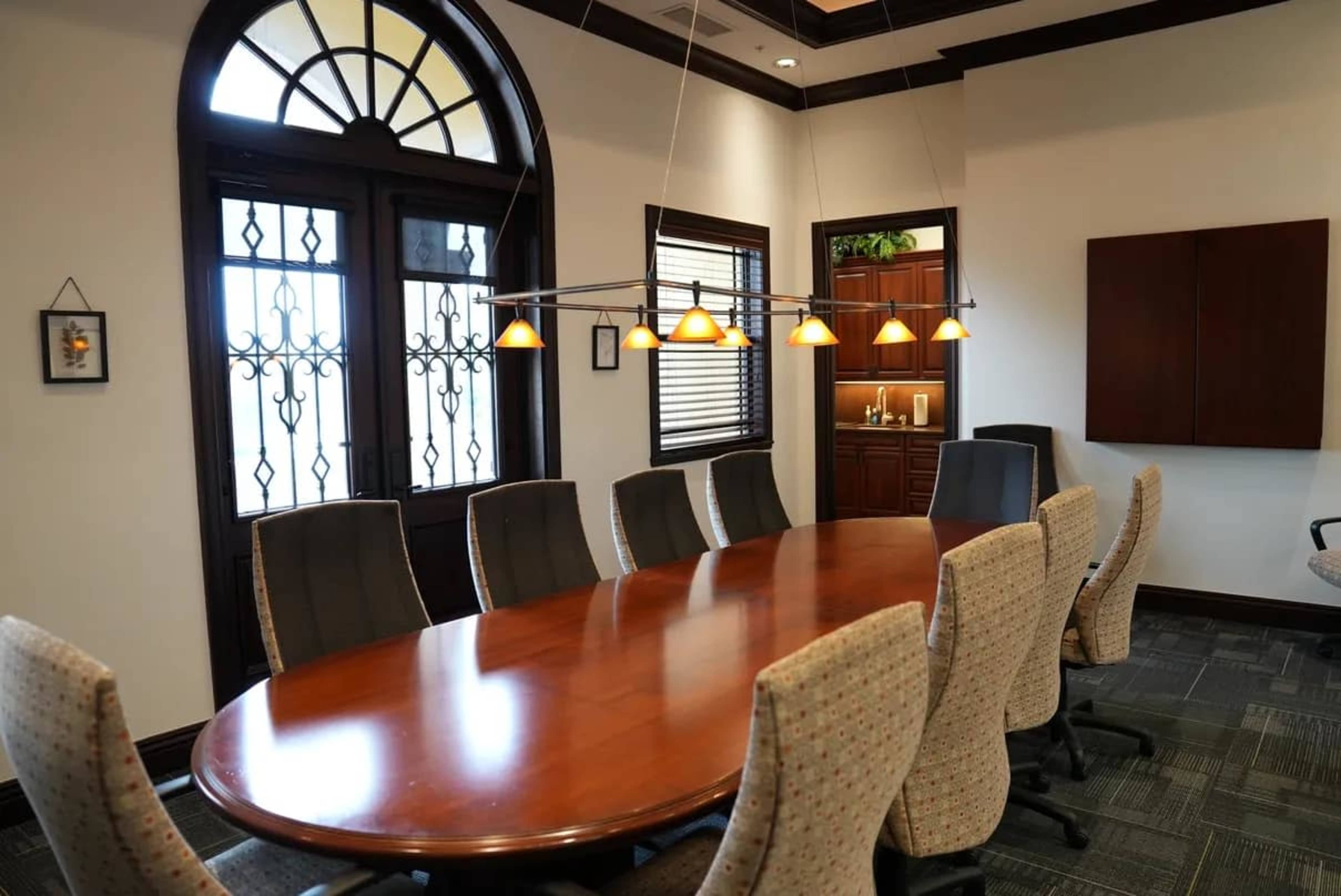 The image shows a conference room featuring an elongated wooden table surrounded by upholstered chairs, with large windows allowing natural light to enter.
