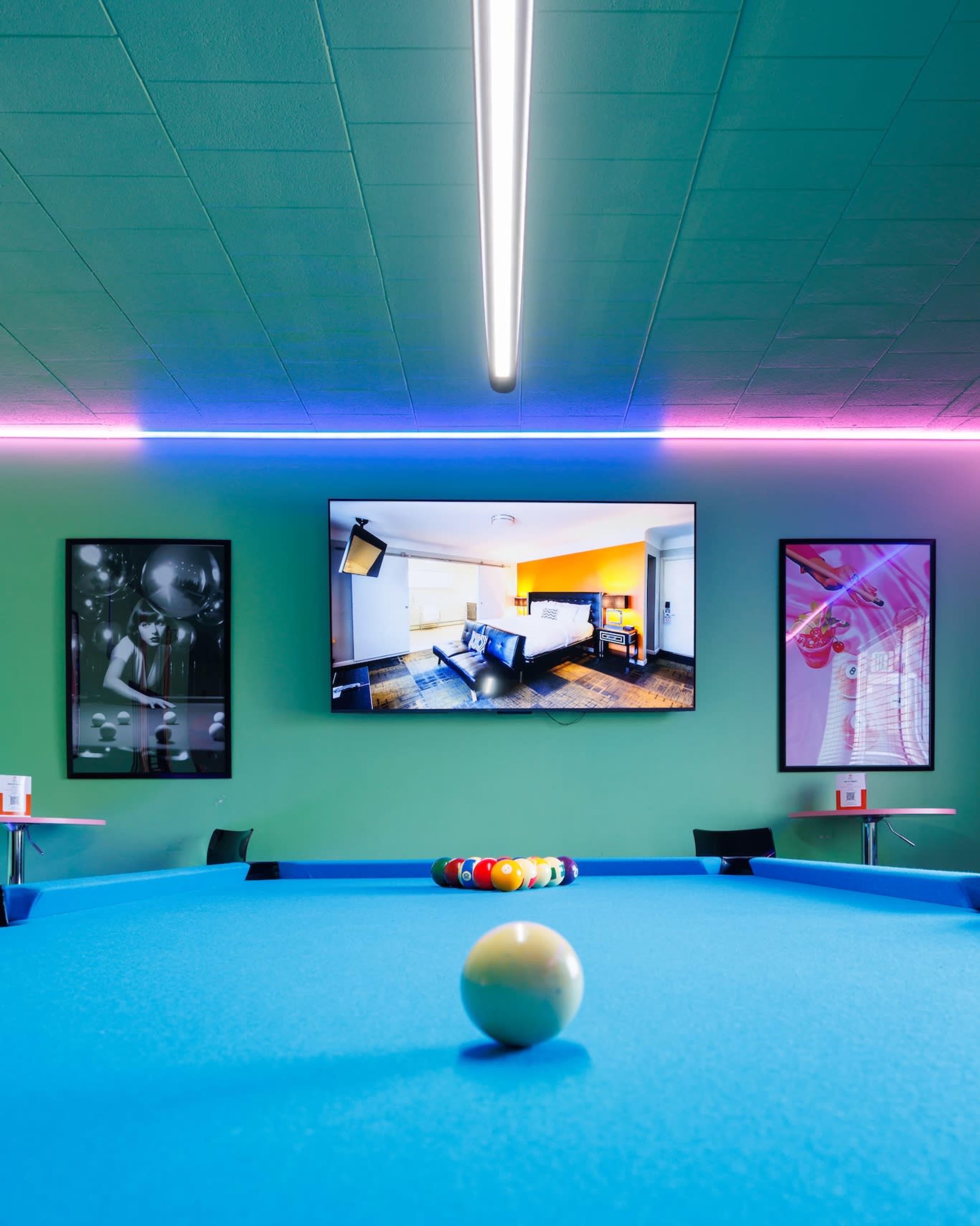 A pool table with a single cue ball sits in front of a colorful room featuring a large television and framed artwork on the walls.