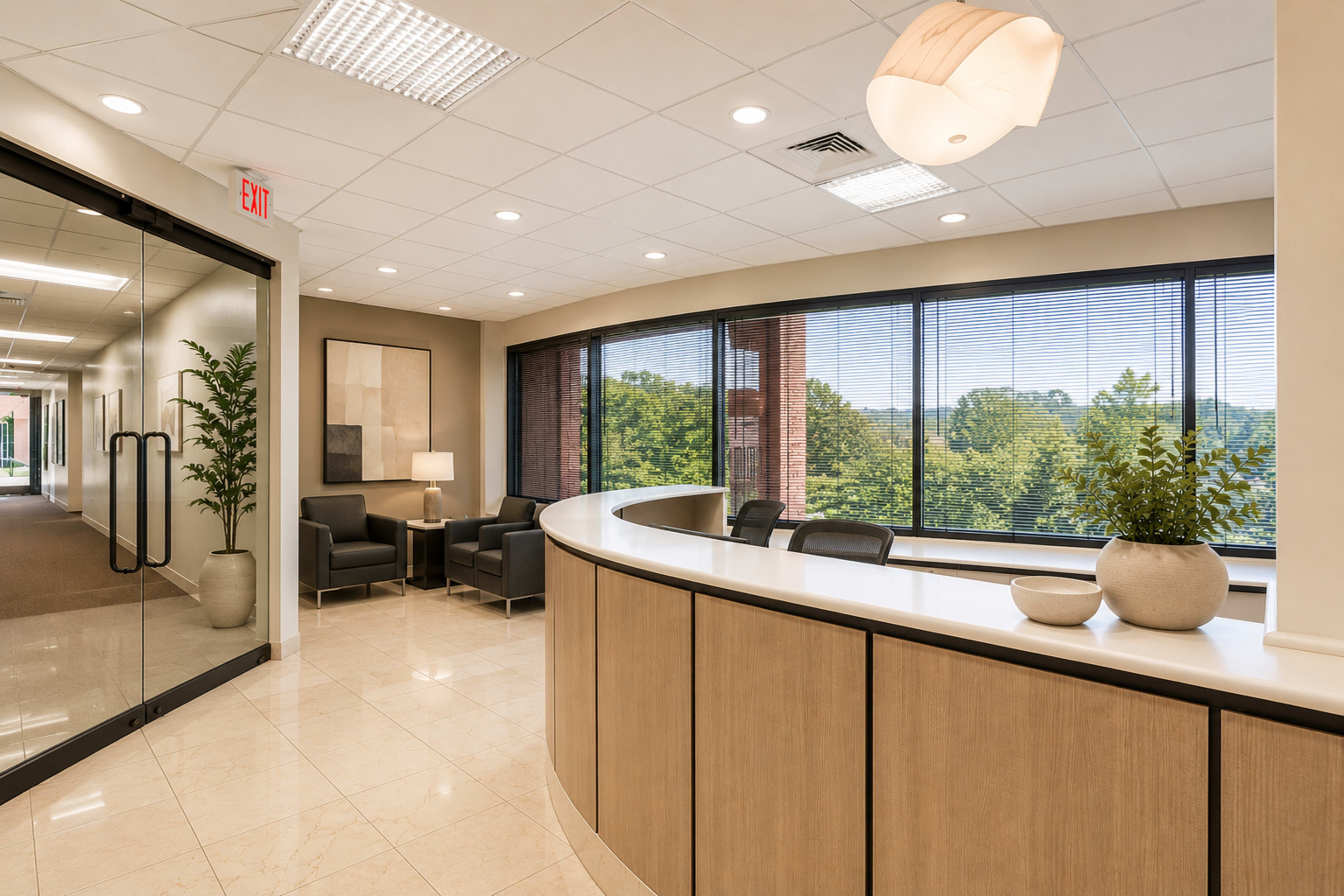 The image shows a modern reception area with a curved front desk, large windows overlooking greenery, and a seating area with two chairs.