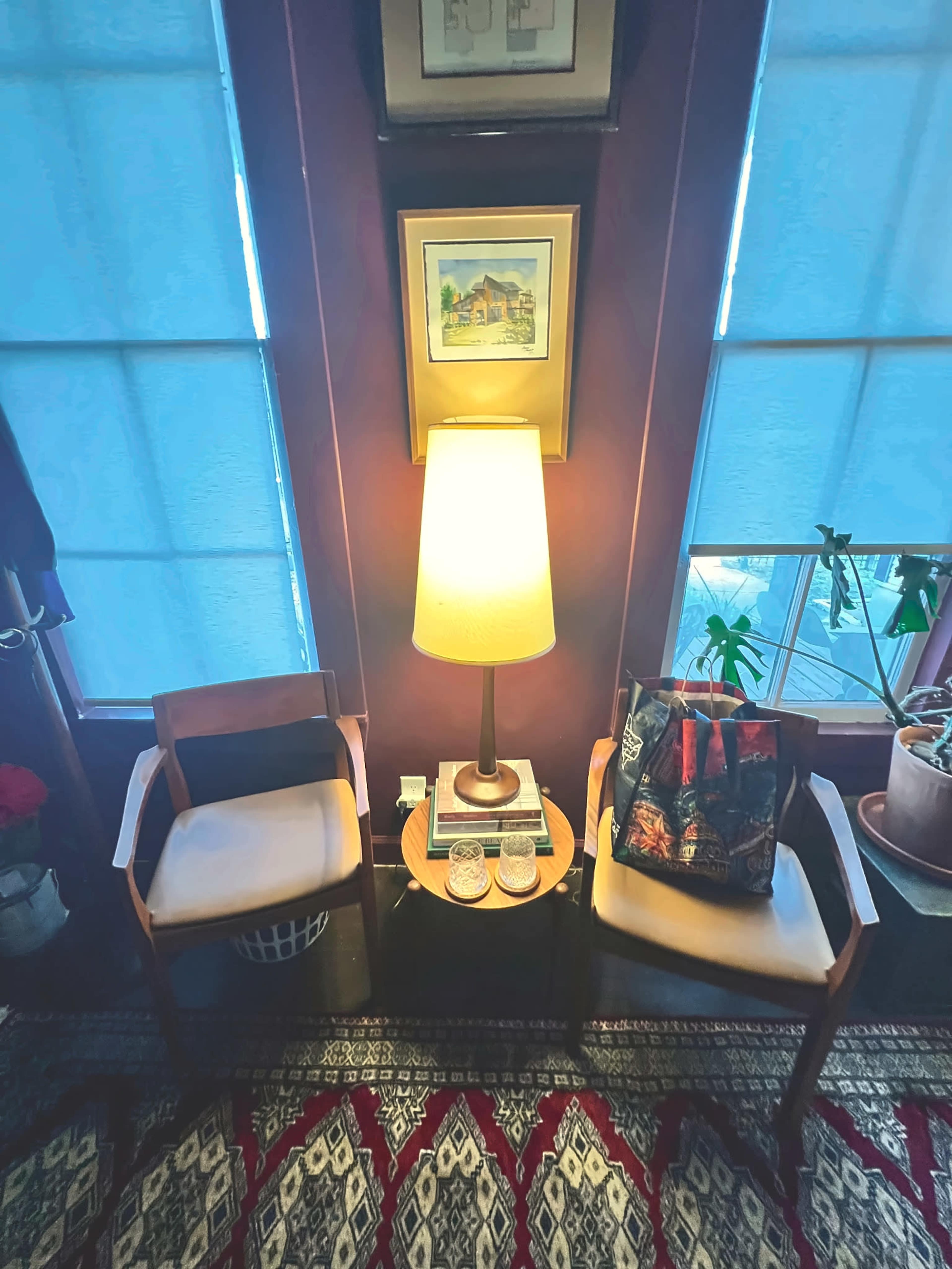 The image shows a corner of a room with two wooden chairs, a small round table holding a lamp and decorative items, and large windows with light filtering through.
