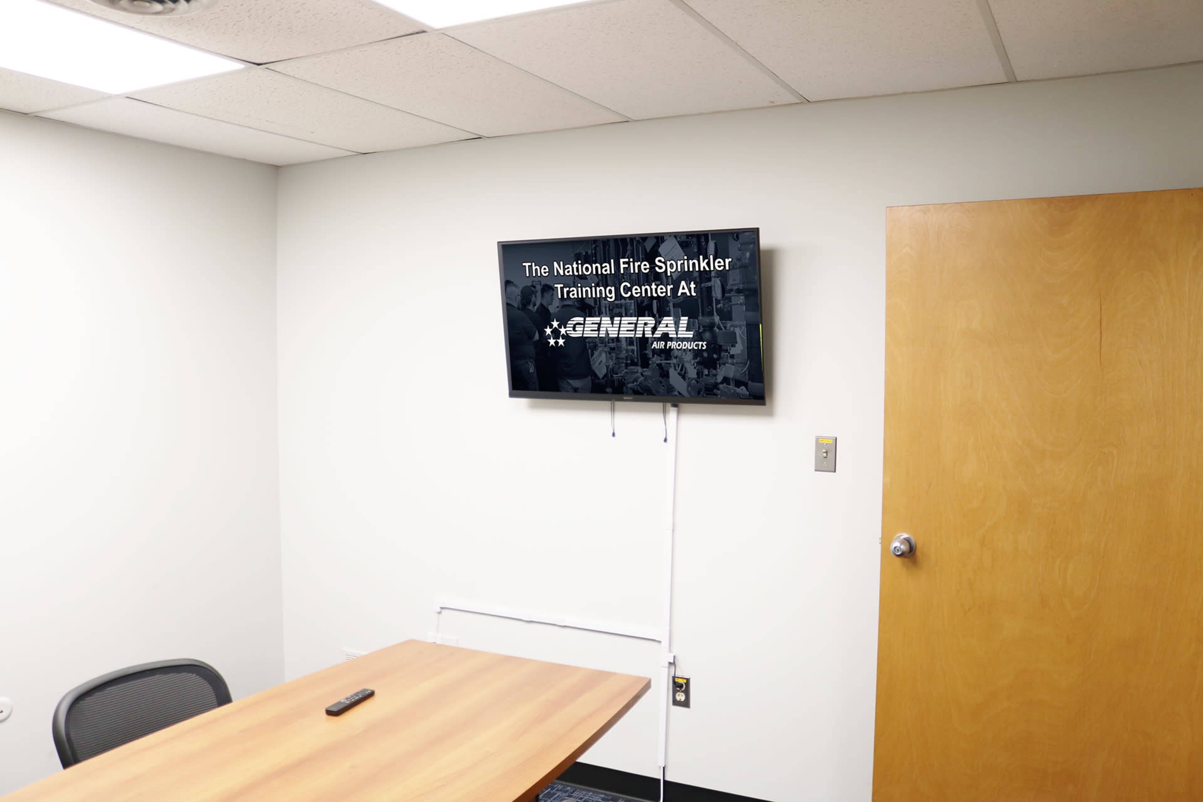 A conference room features a wall-mounted television displaying text about the National Fire Sprinkler Training Center at General Fire Products, alongside a wooden table and a chair.