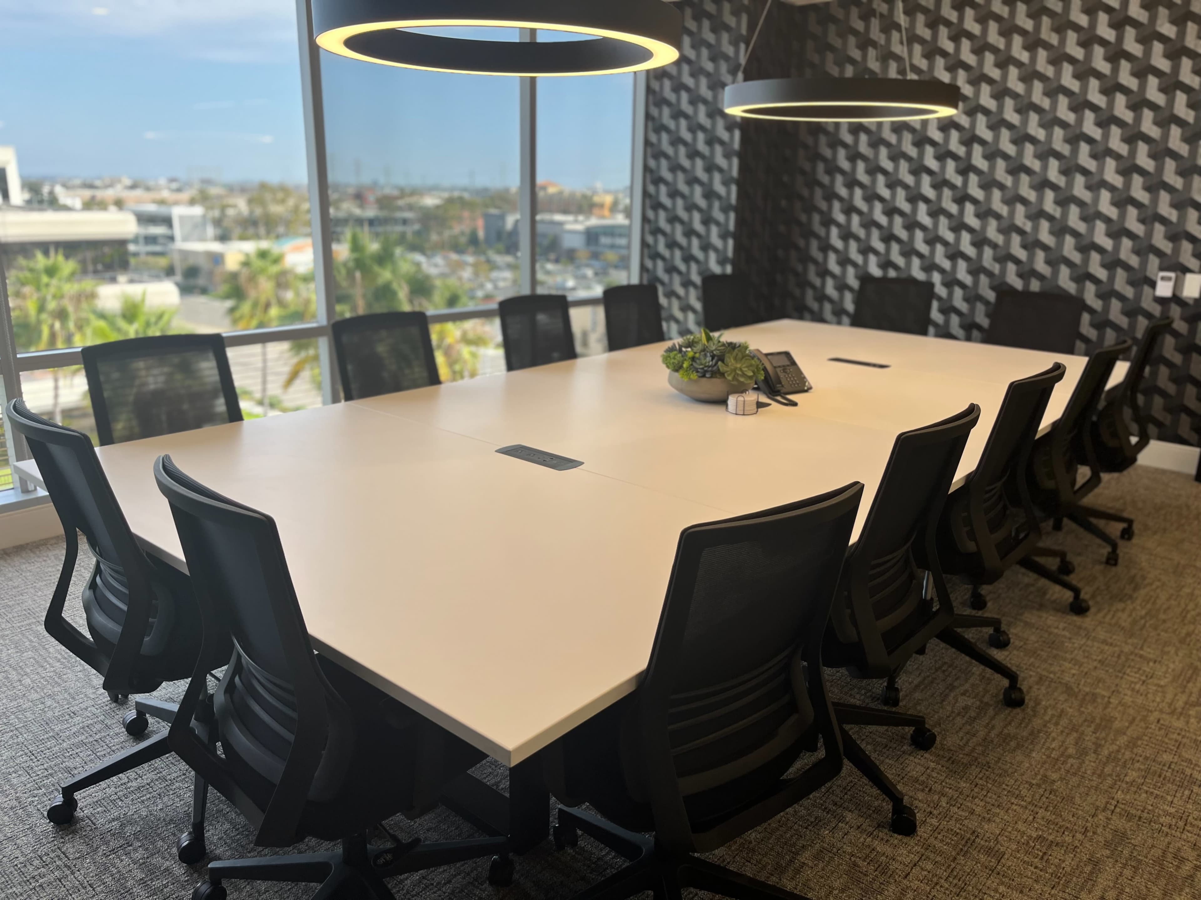 A modern conference room features a large rectangular table surrounded by ergonomic chairs, with two pendant lights overhead and a view of the city through large windows.