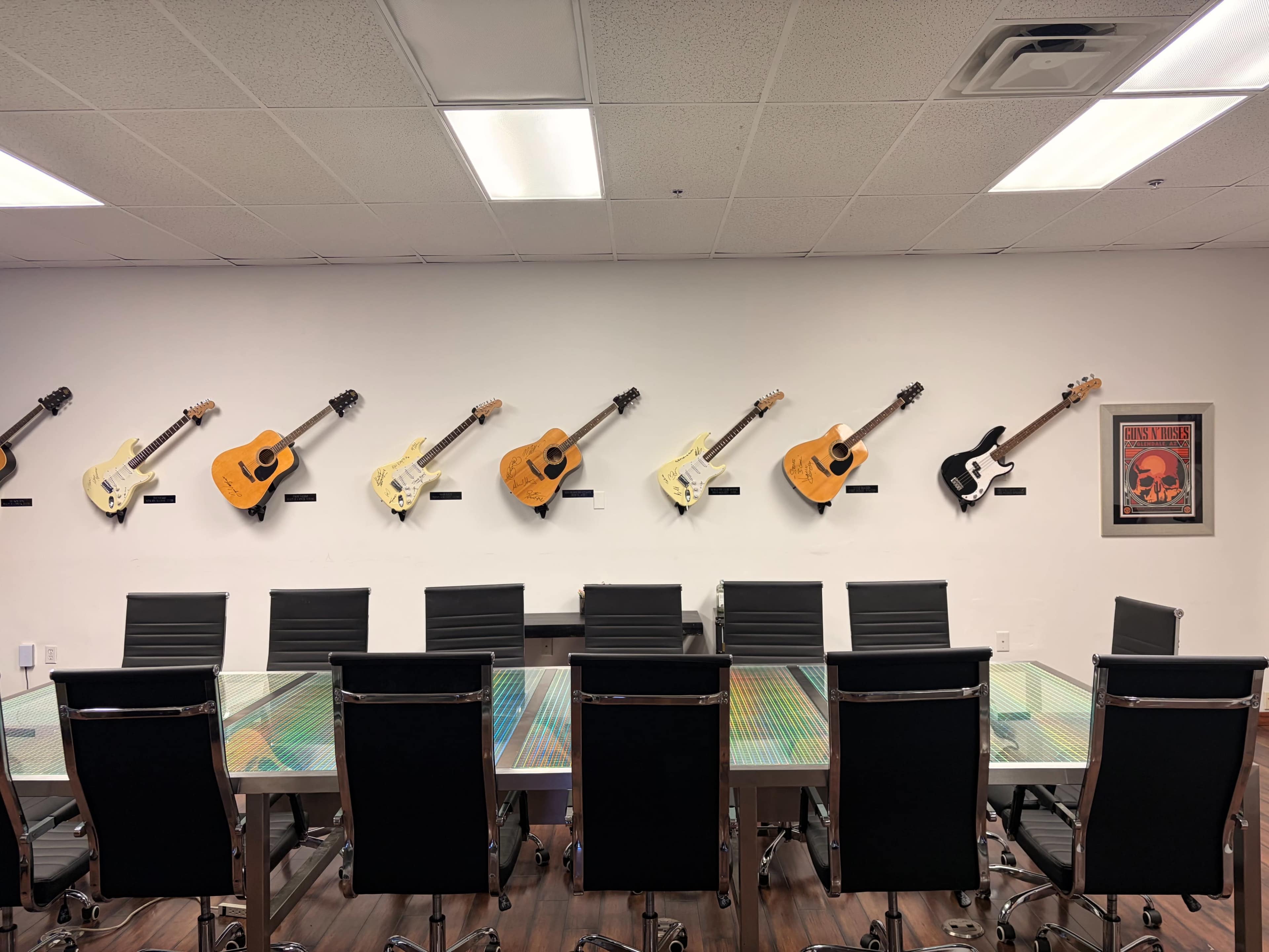 A conference room features a long glass table with black chairs and a wall displaying several guitars alongside a framed poster.