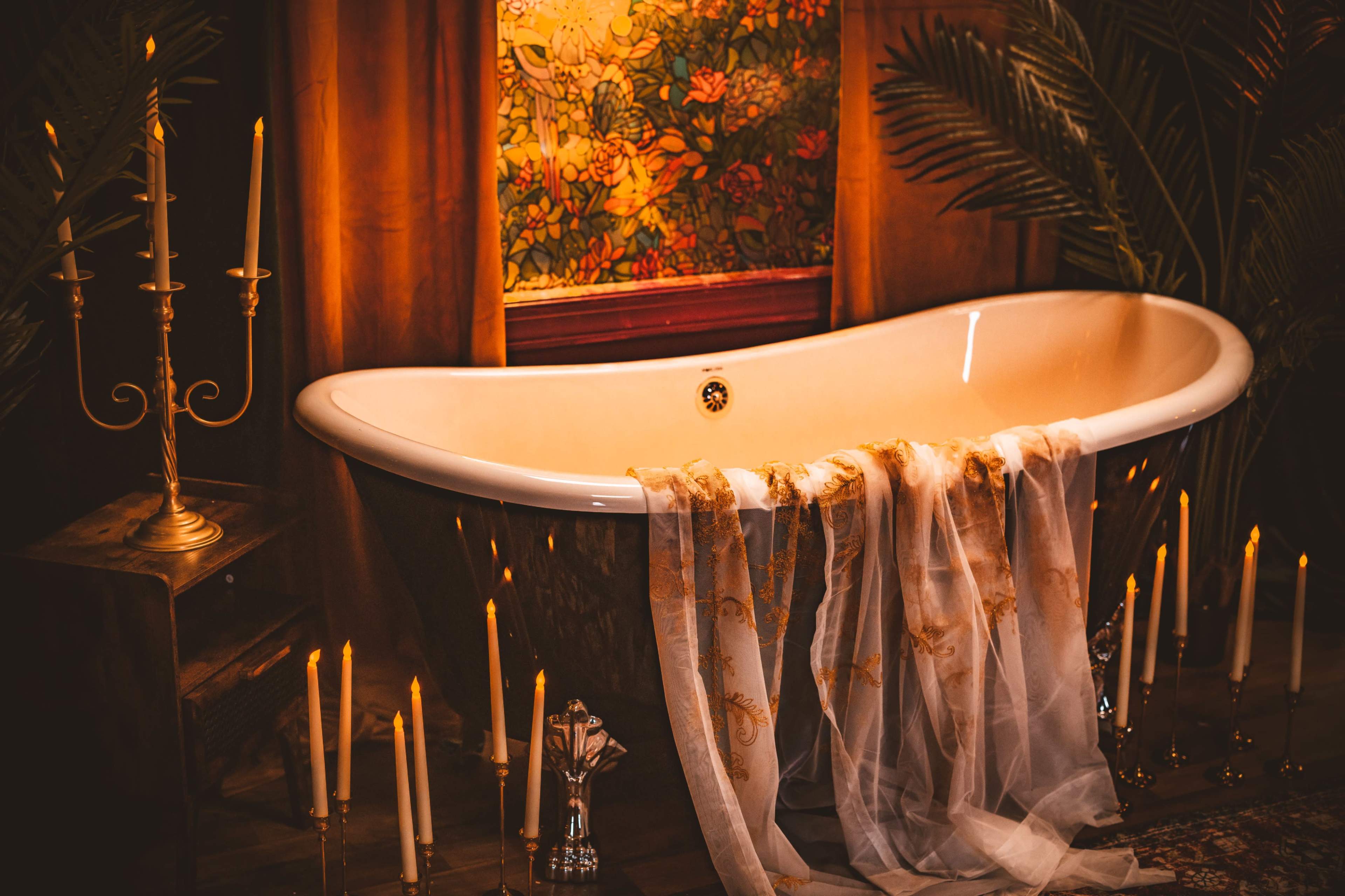 A vintage clawfoot bathtub is adorned with flowing fabric and surrounded by candles and lush greenery in a softly lit setting.