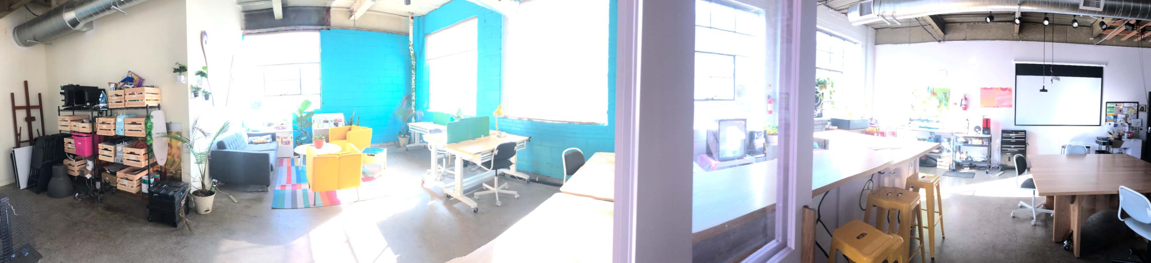 The image shows a bright, open workspace with various seating areas, desks, and plants, featuring a blue accent wall and large windows.