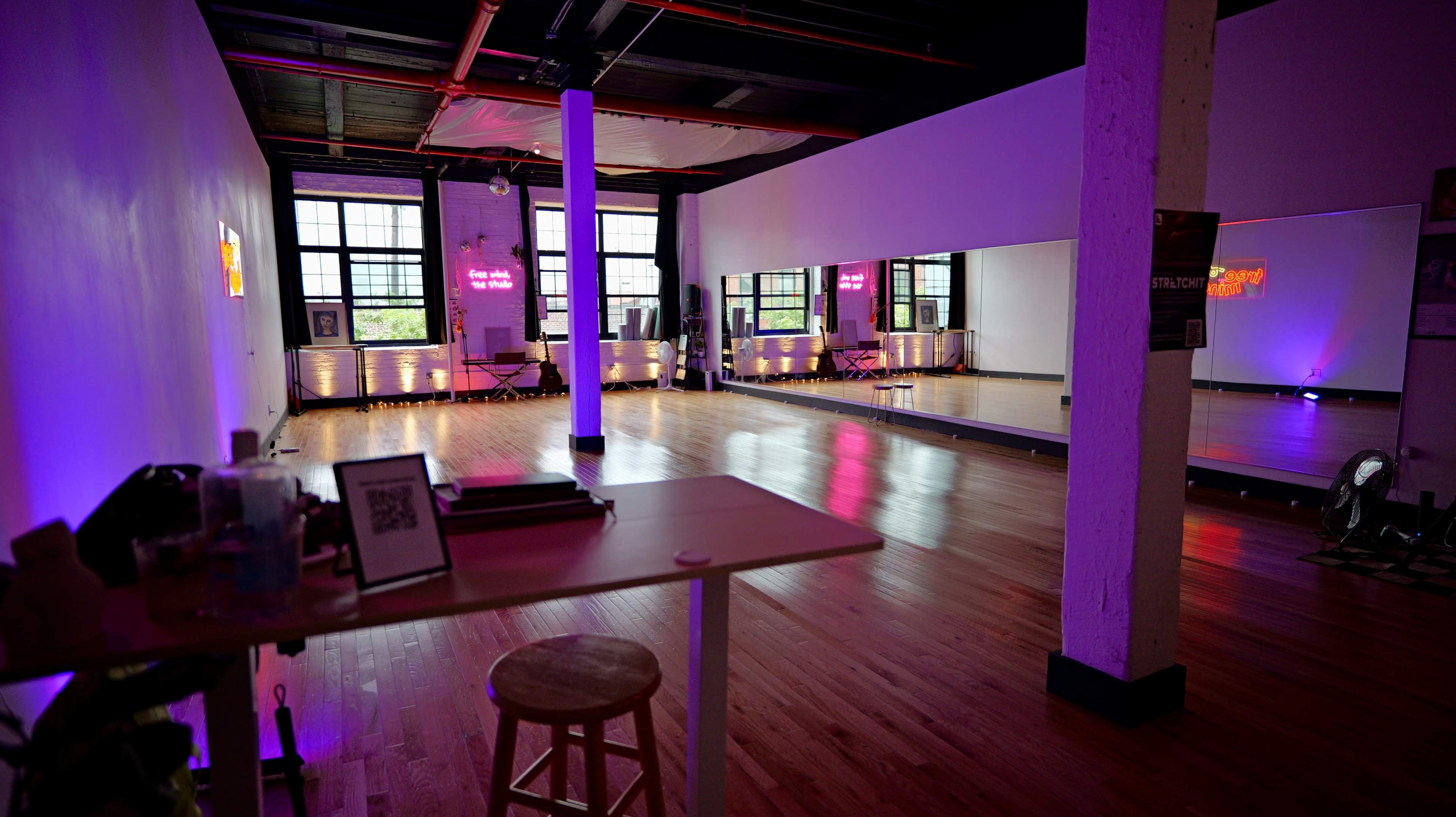 An empty dance studio with wooden floors, large mirrors, and purple lighting, featuring a small table and stool in the foreground.