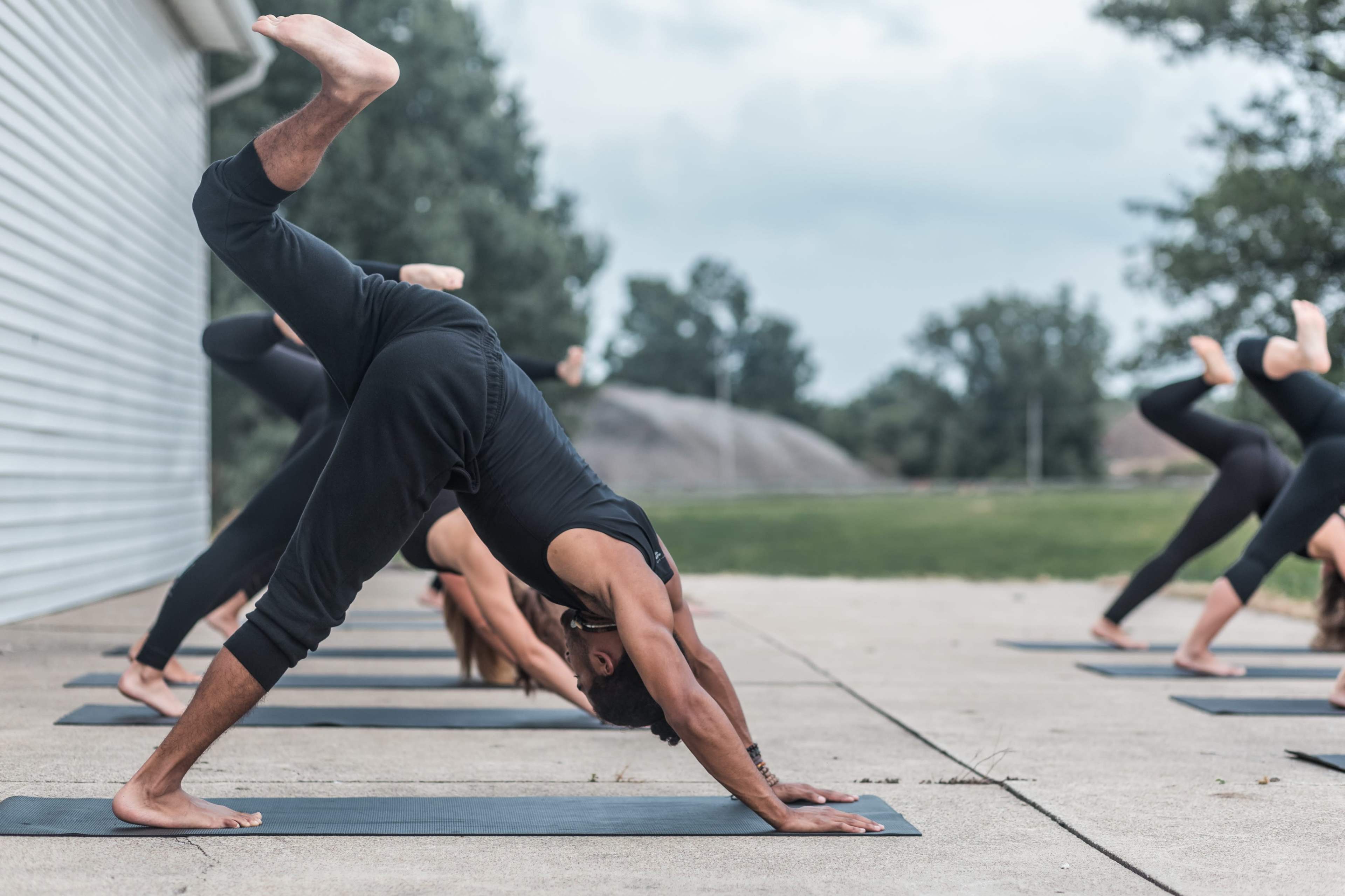 A group of individuals practice yoga outdoors on mats, with one person in a downward facing dog pose.