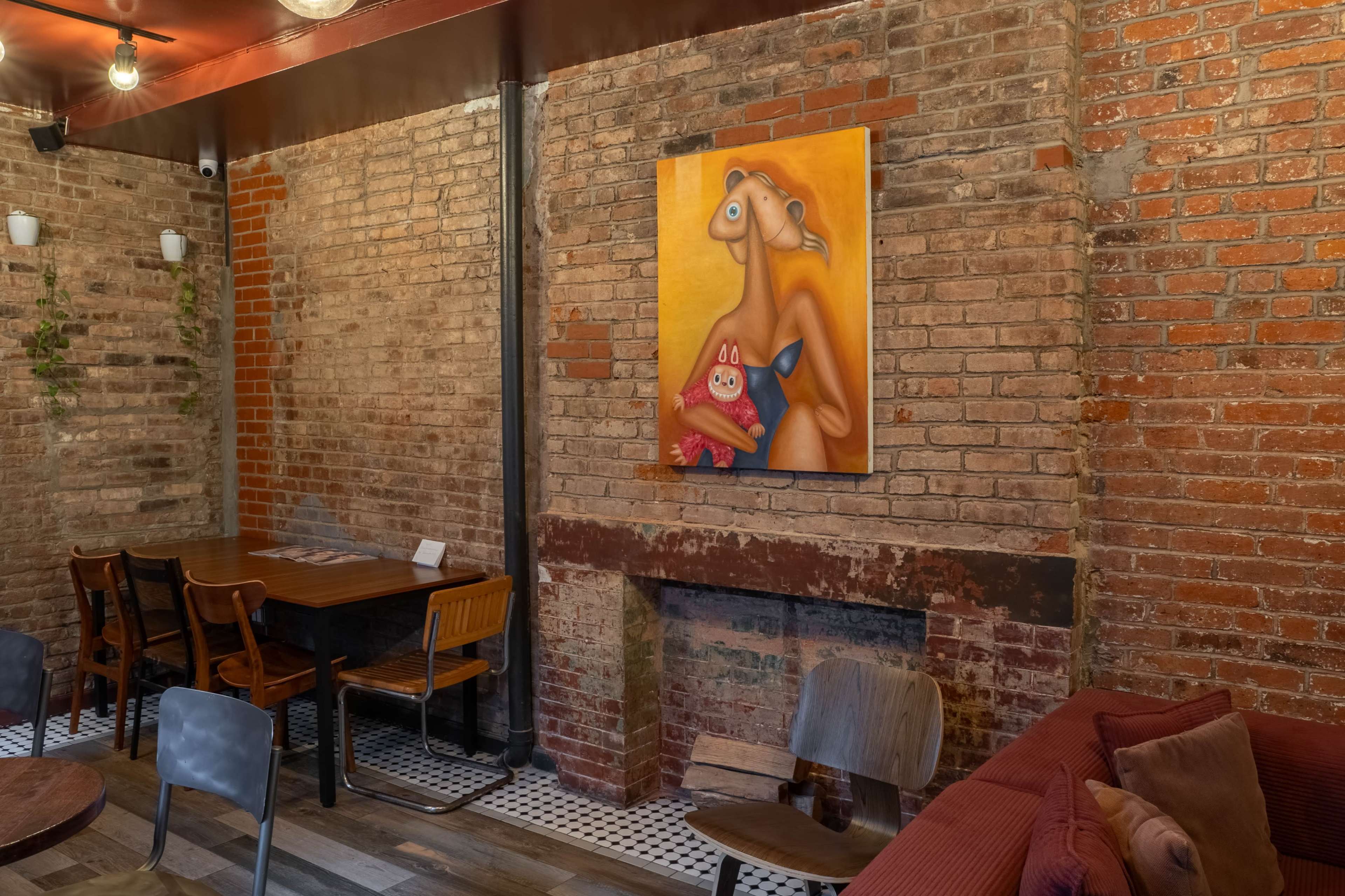 The image shows a cozy interior of a restaurant or café with exposed brick walls, a wooden table surrounded by chairs, and a colorful painting hanging above a brick fireplace.