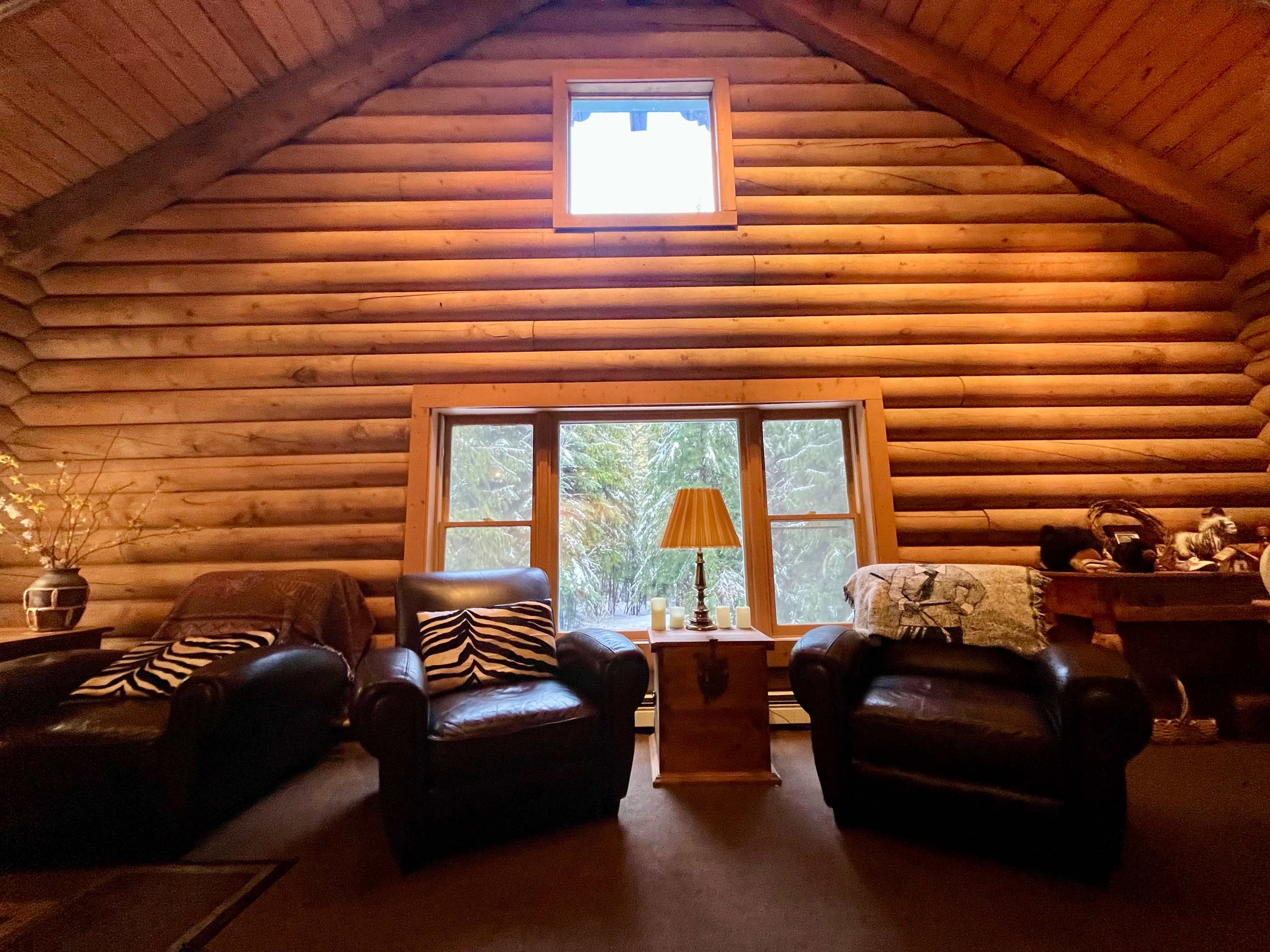 A cozy room with two leather armchairs, a side table with a lamp, and wooden walls illuminated by natural light from a window.