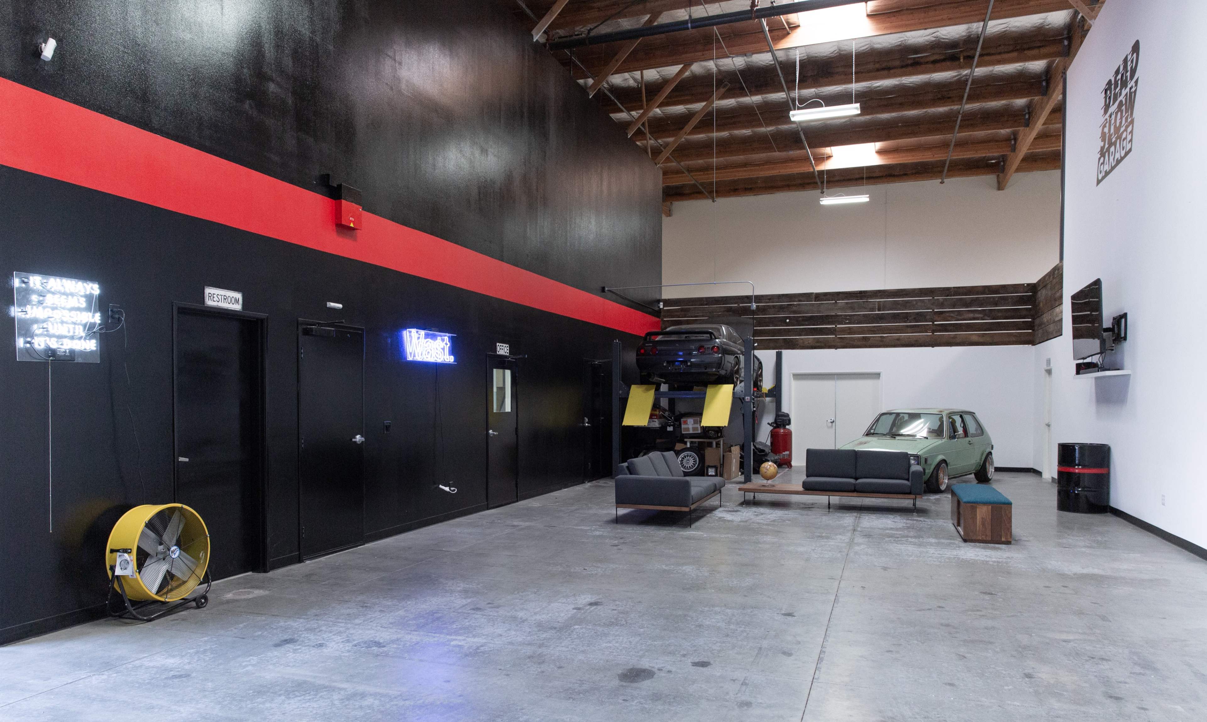 A spacious, minimalistic interior of a garage or warehouse with a high ceiling, featuring a parked vehicle, a modern sofa, and neon signage on the walls.