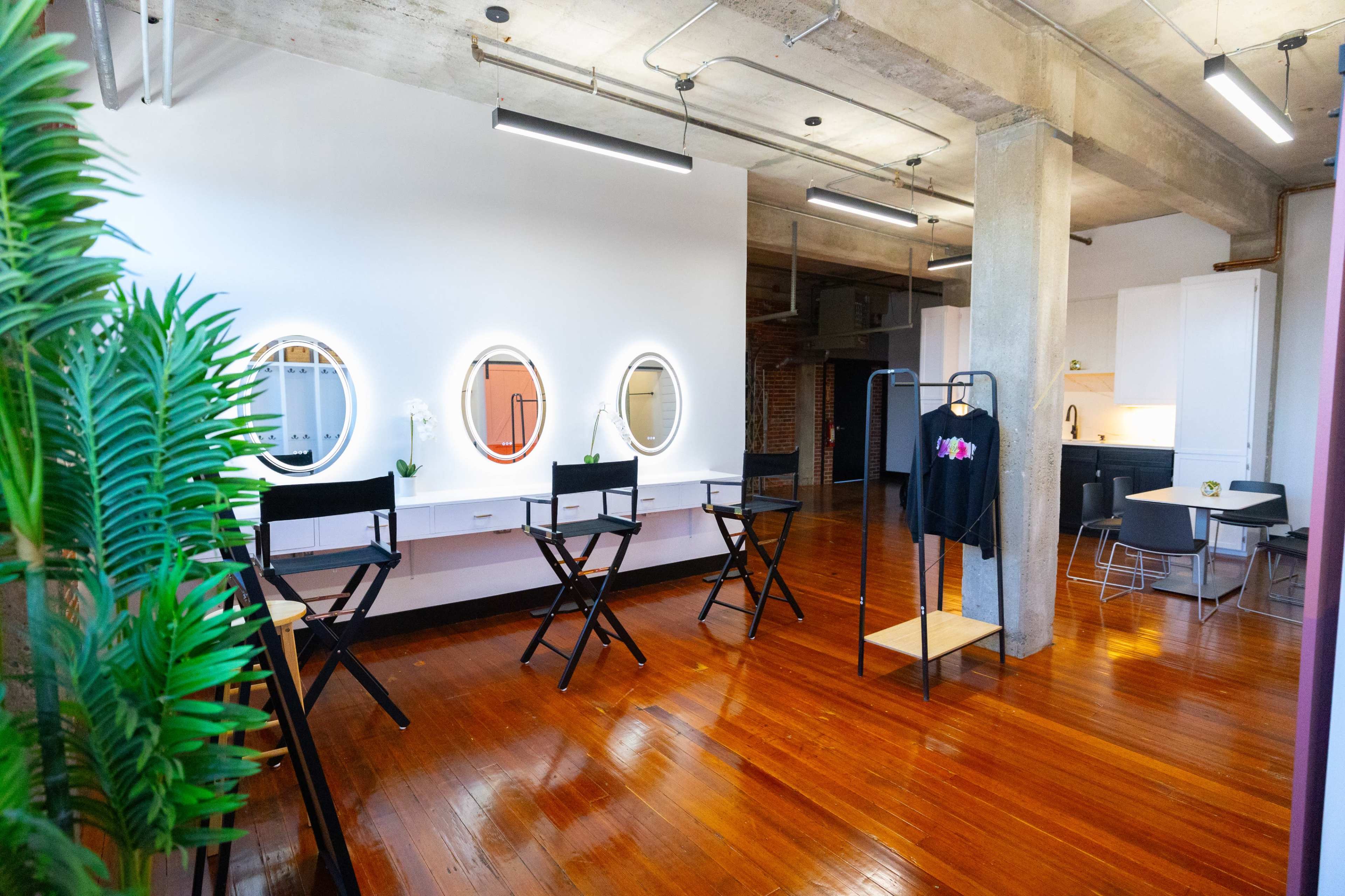 A spacious studio with wooden floors, equipped with makeup stations featuring circular mirrors and director chairs, along with a clothing rack and a small kitchenette in the background.