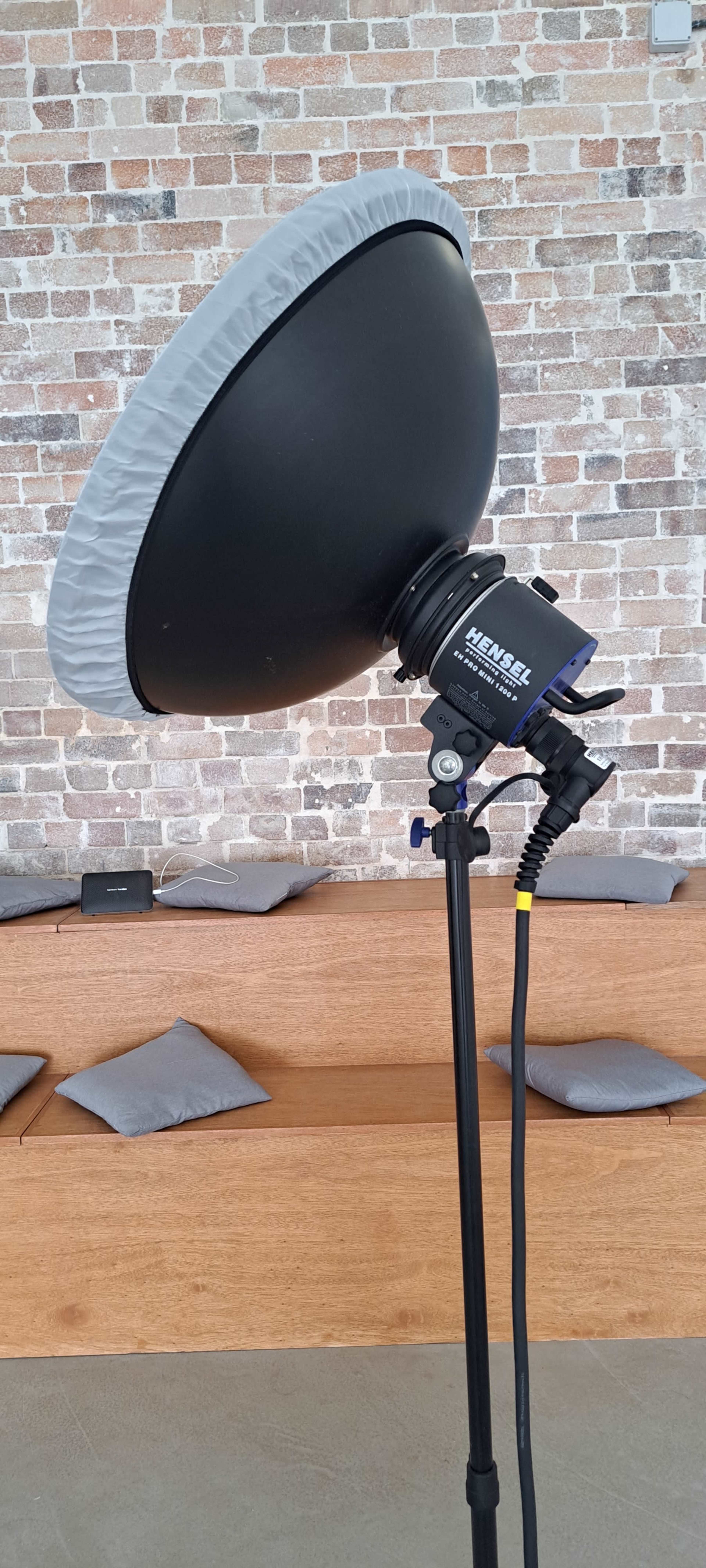 A large, round softbox light is mounted on a stand in front of a brick wall, with wooden benches and cushions visible in the background.