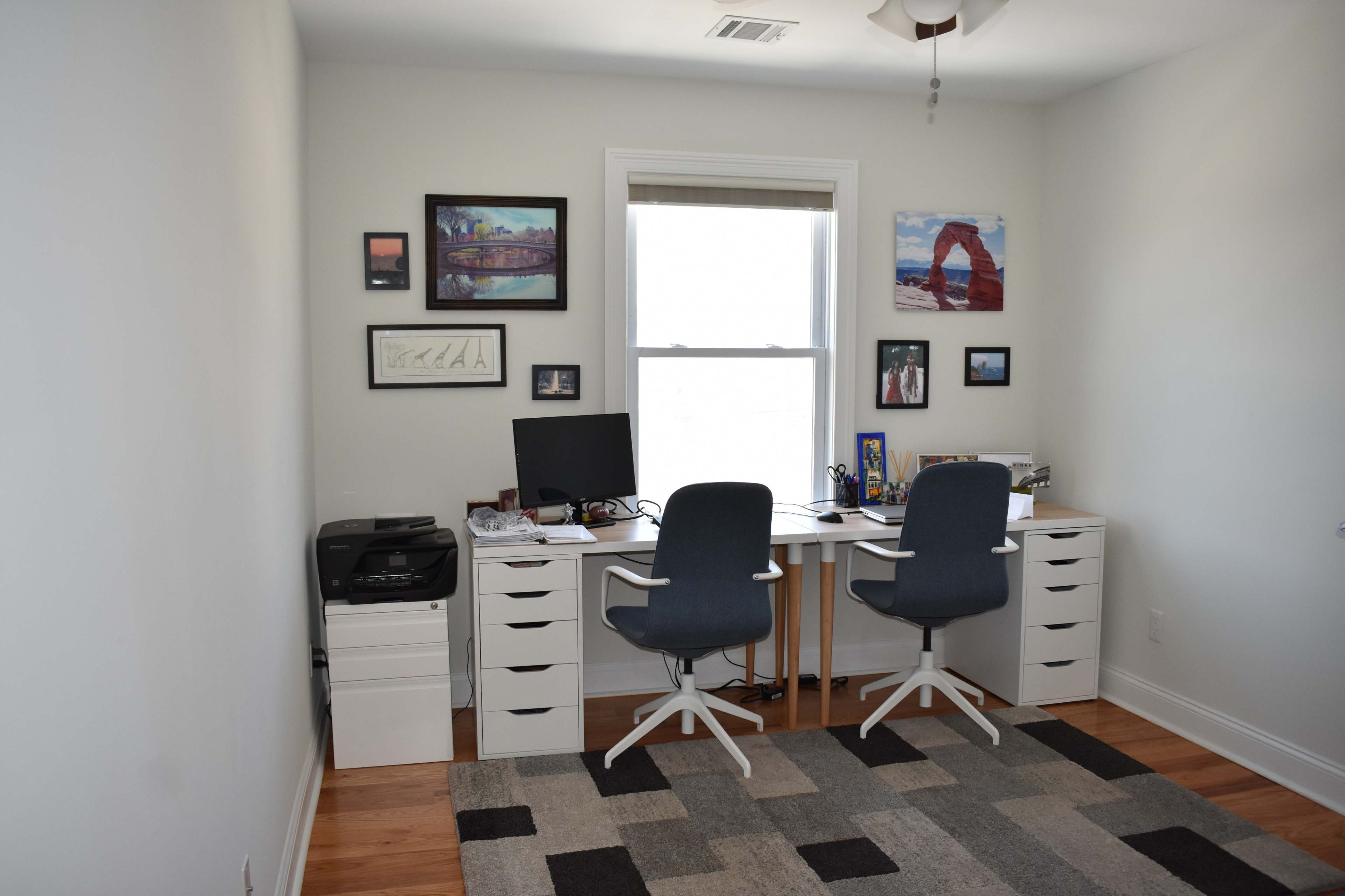 A well-organized home office features two chairs at a desk with a computer monitor, printer, and framed artwork on the walls.