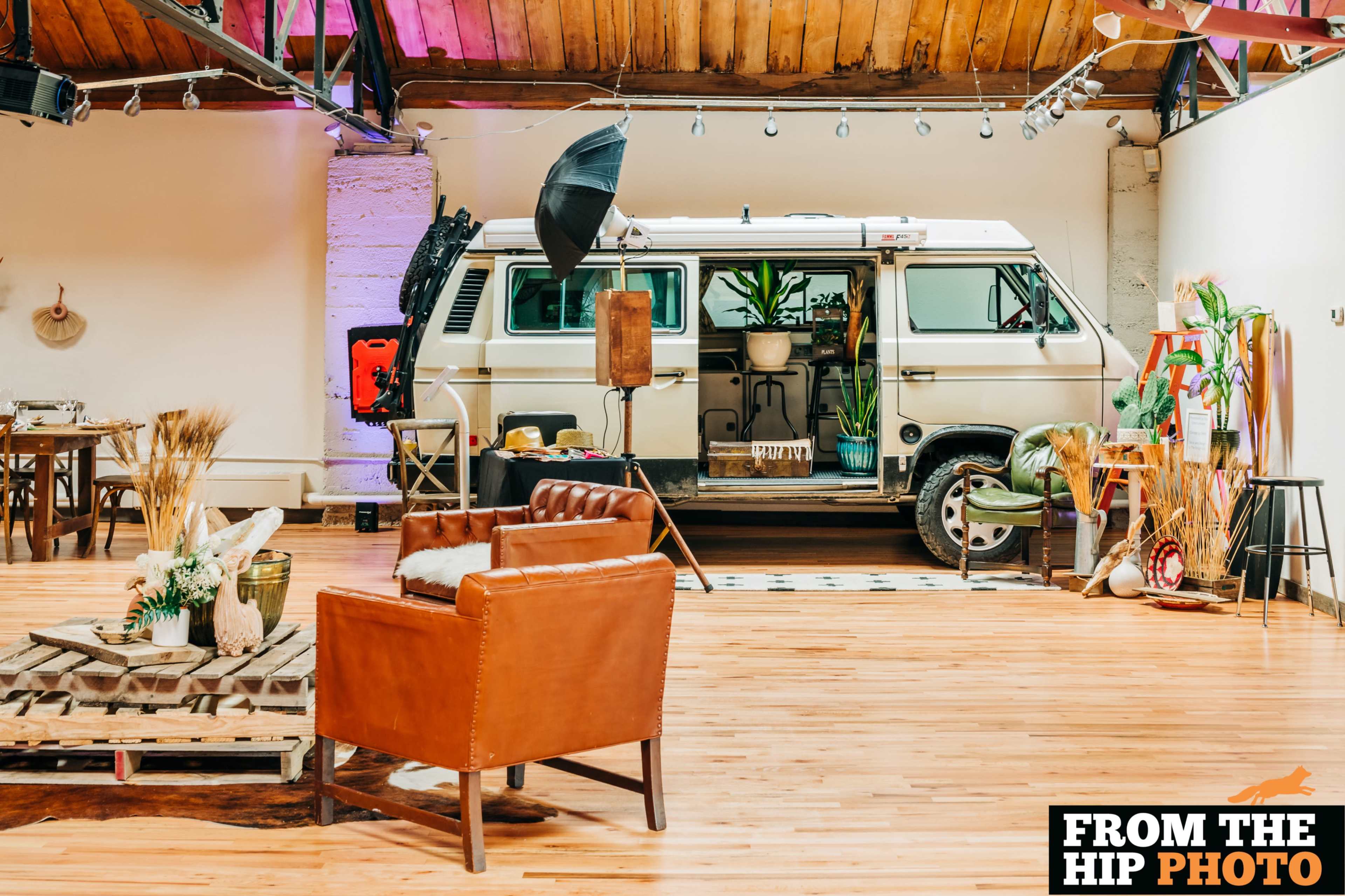 The image shows a spacious indoor setting featuring a vintage van parked against a wall, surrounded by rustic furniture and decorations.