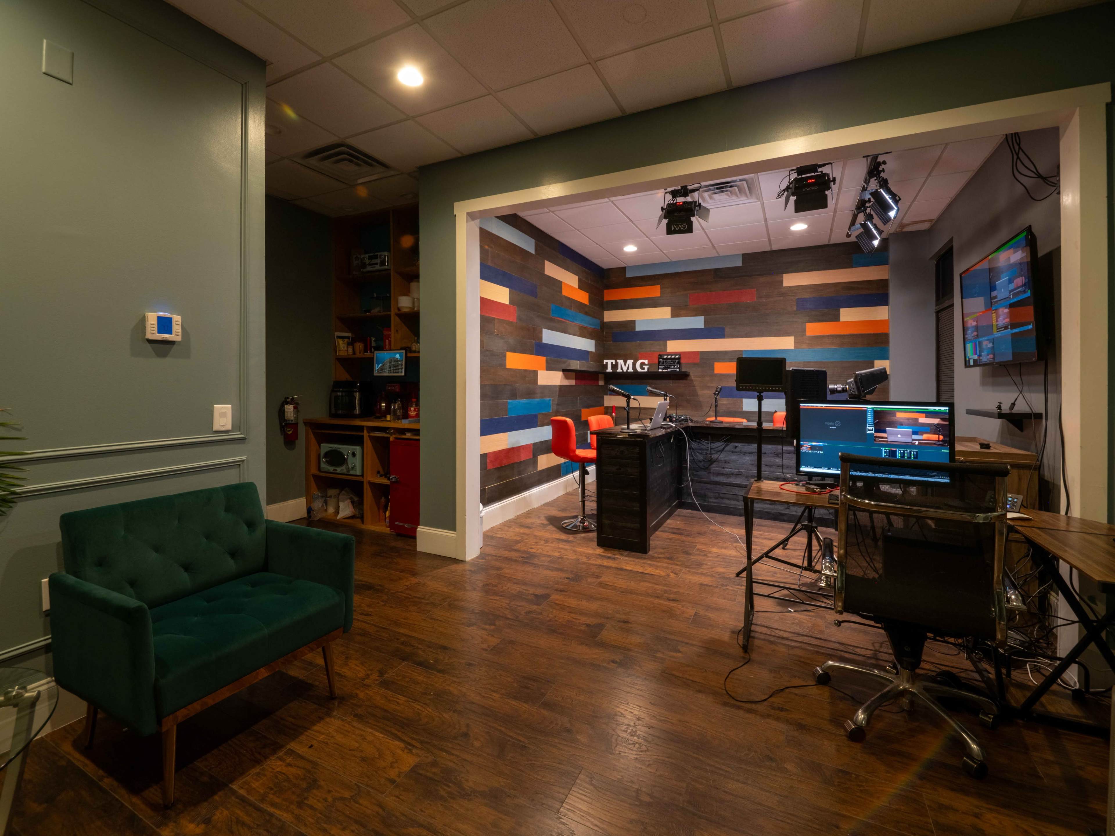 A modern office space featuring a studio setup with colorful wall panels, a desk with multiple monitors, and a lounge area with a green upholstered sofa.