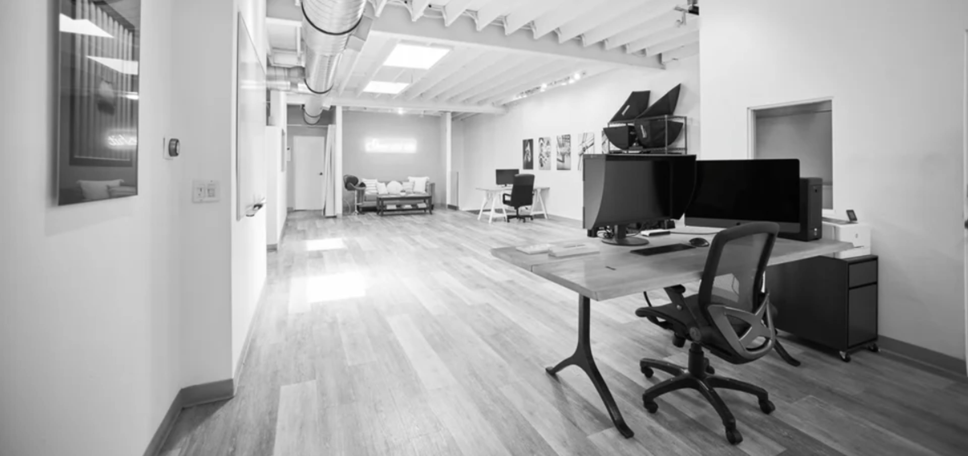 The image shows a spacious, modern office interior featuring wooden flooring, a desk with dual monitors, and various seating areas.