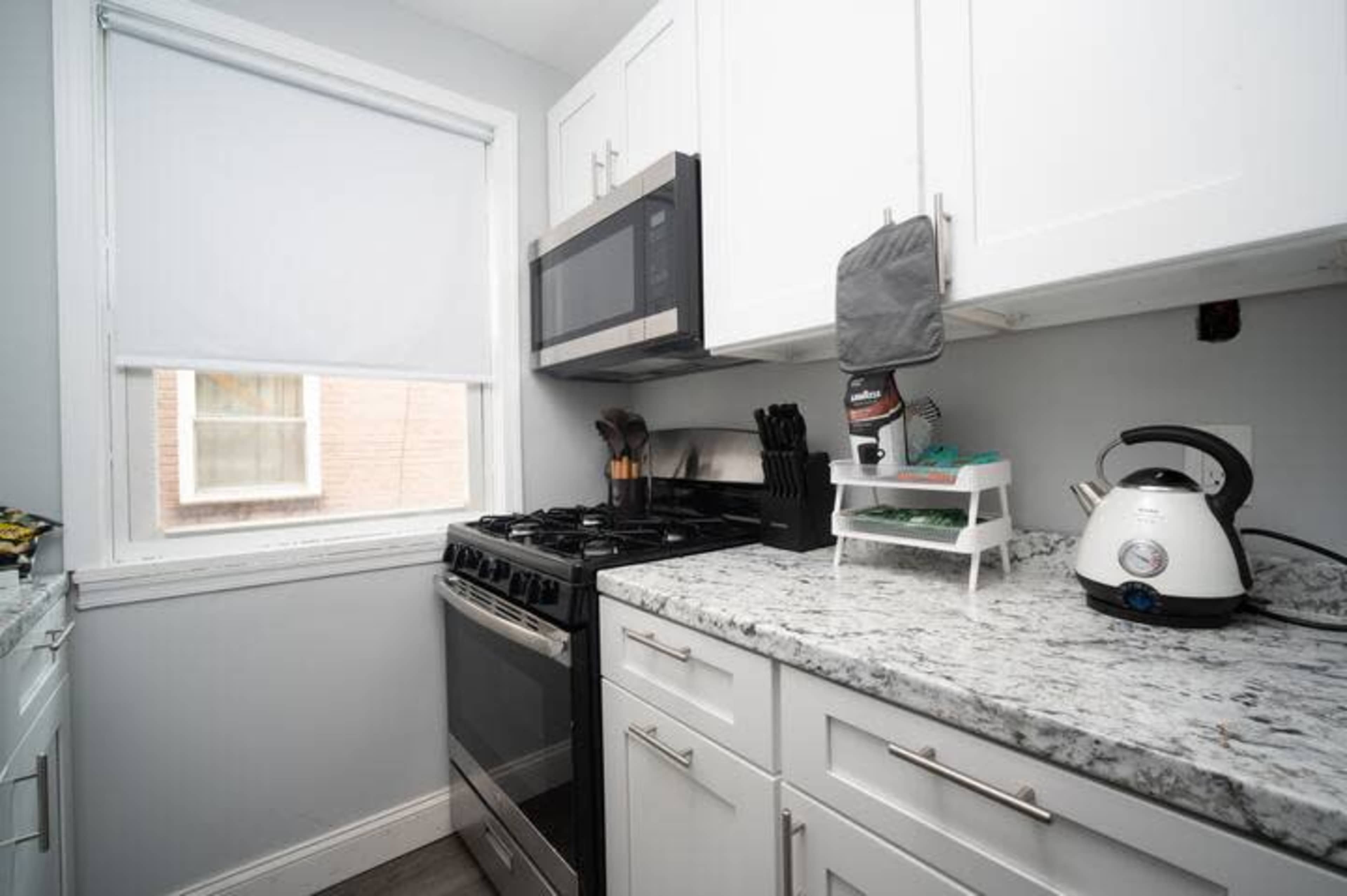 A compact kitchen with white cabinets, a stainless steel microwave, and a granite countertop with a gas stove and kettle.