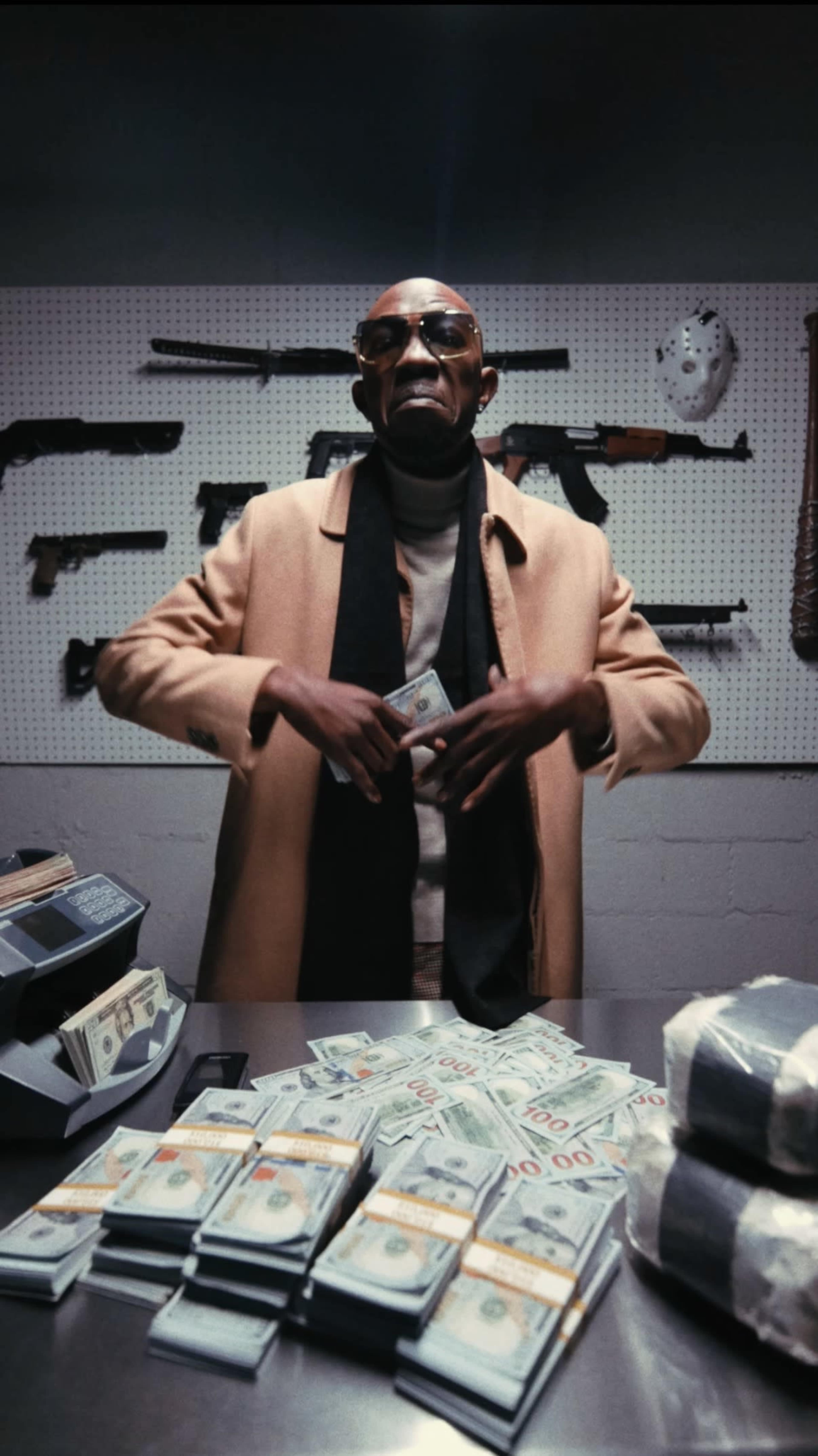 A man in sunglasses stands at a table covered with stacks of cash, with firearms displayed on a wall behind him.
