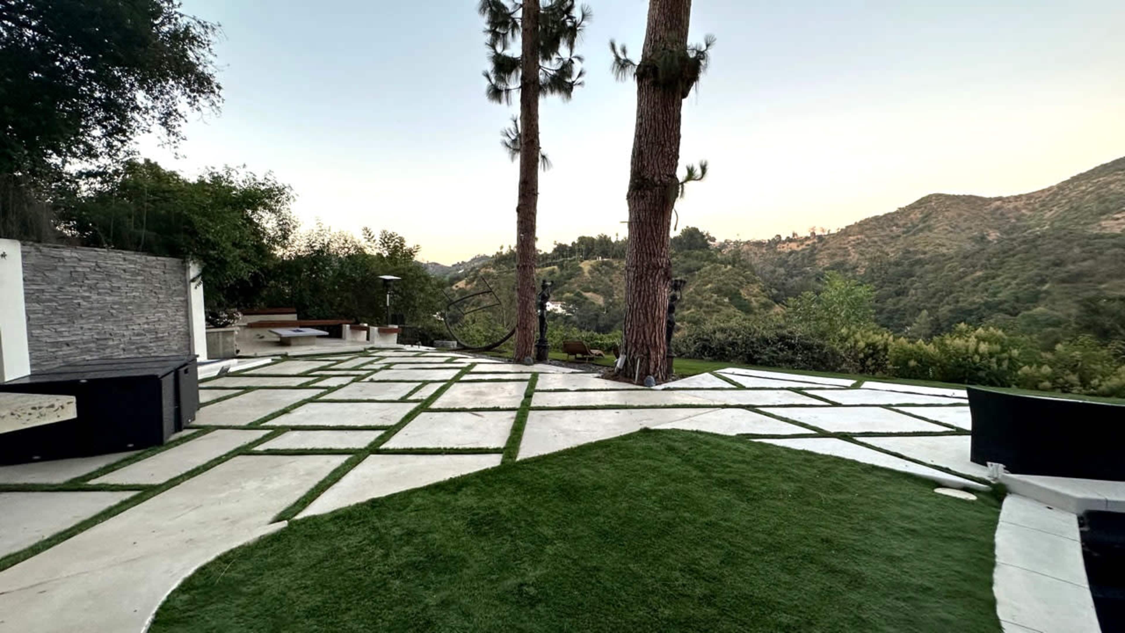 A landscaped outdoor area with geometric patterns of concrete and grass, bordered by two tall trees and a view of a hilly landscape in the background.