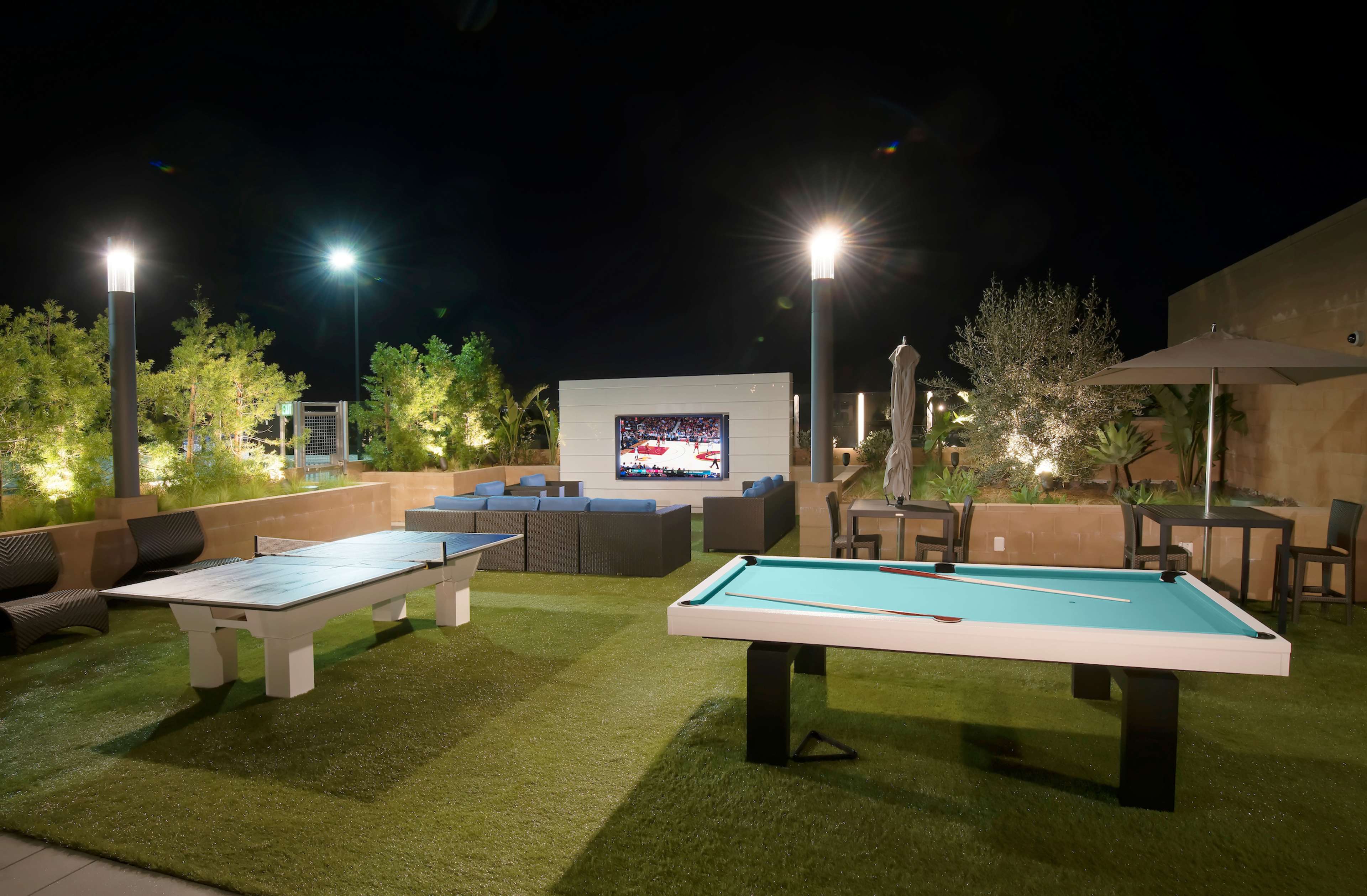 A rooftop patio at night featuring a pool table, outdoor seating, and a large screen displaying a sports event.