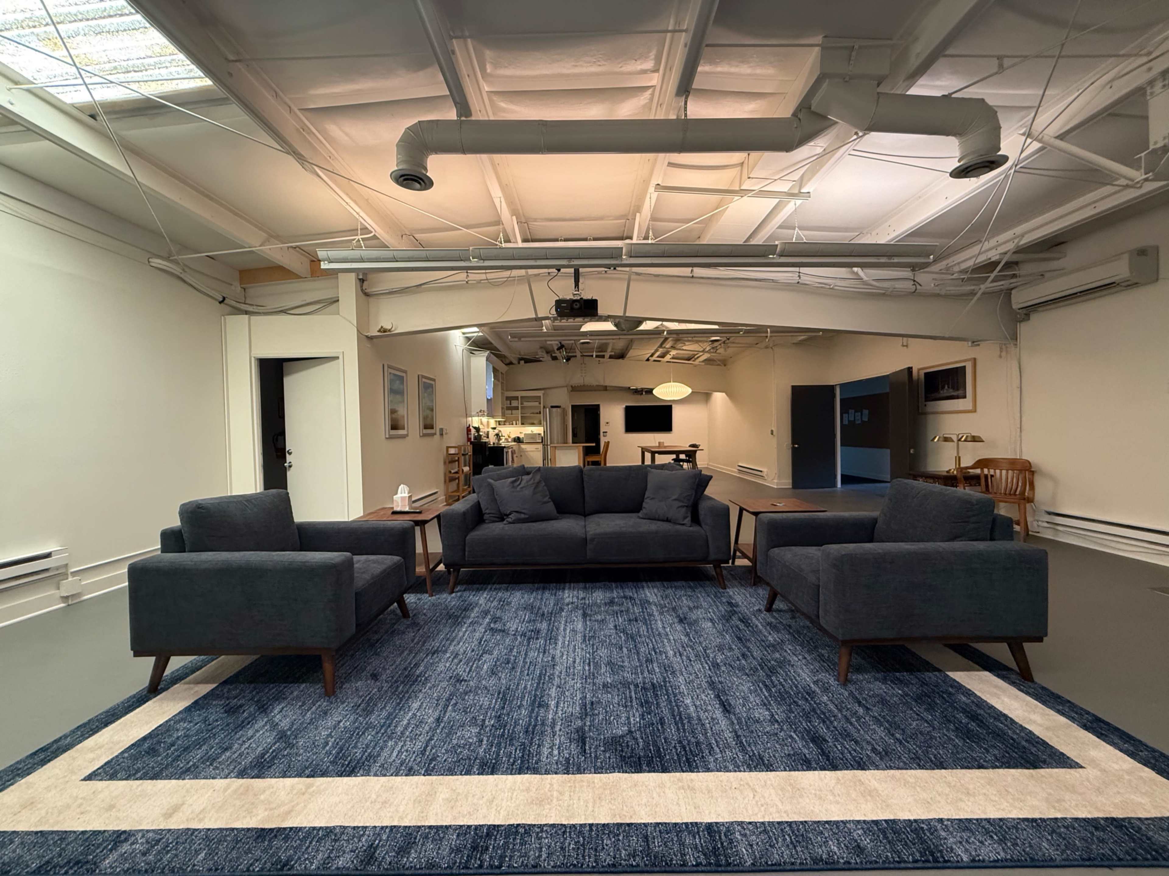 The image shows a spacious, minimalistic room furnished with a dark sectional sofa, two matching armchairs, and a large blue area rug.