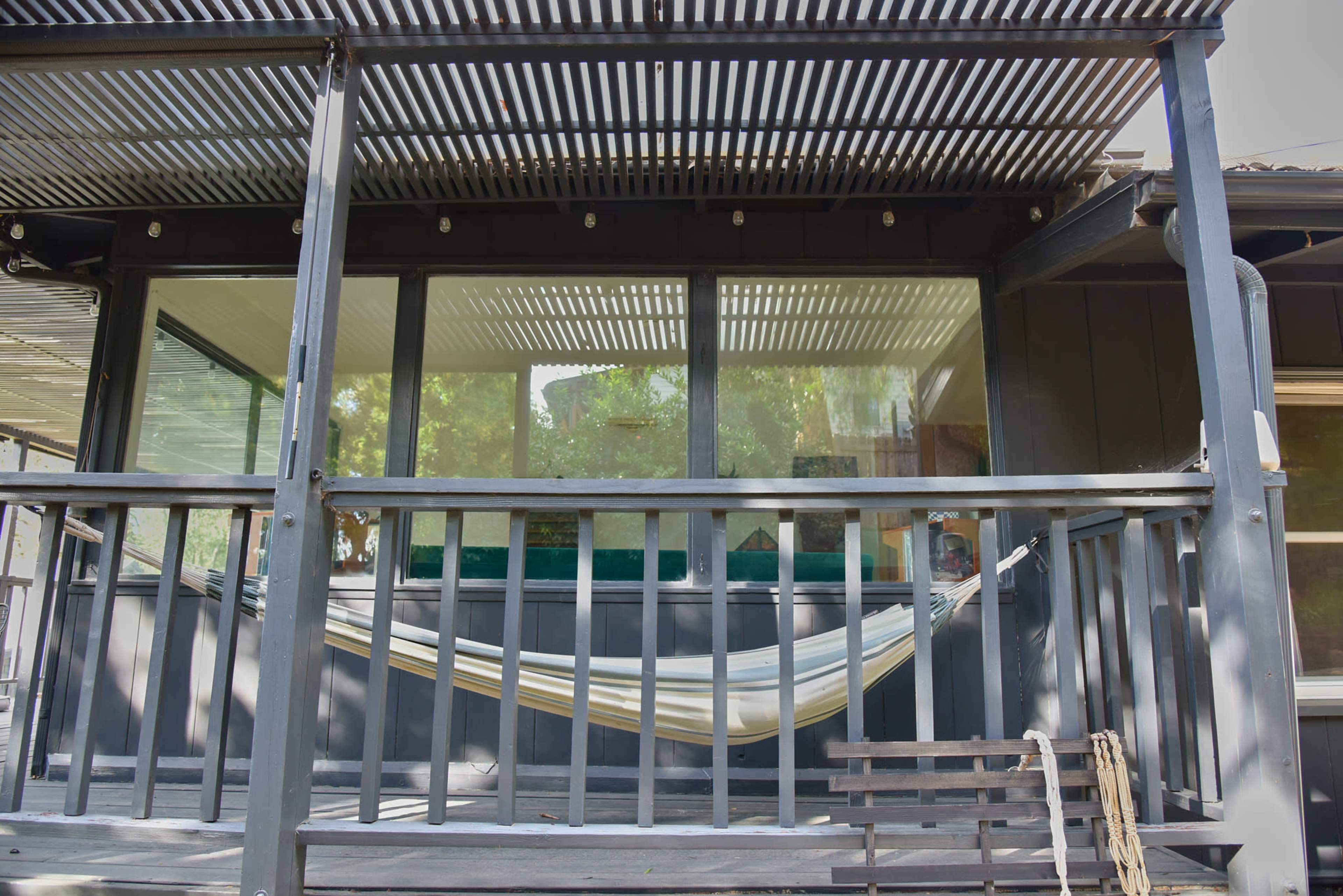A porch with a hammock hanging between two posts, beneath a slatted roof structure and a large window.