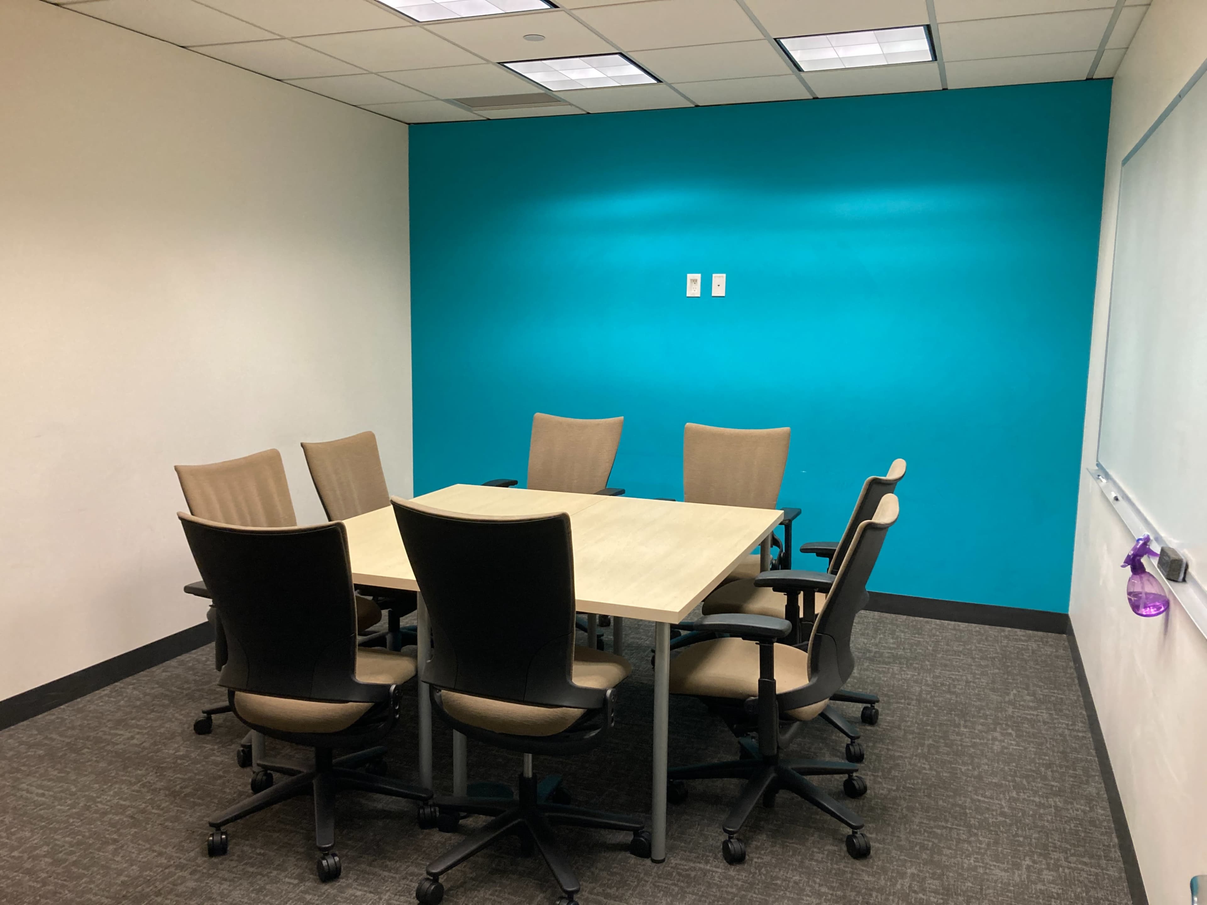 A conference room features a rectangular table surrounded by six chairs, with a bright turquoise wall and a whiteboard.