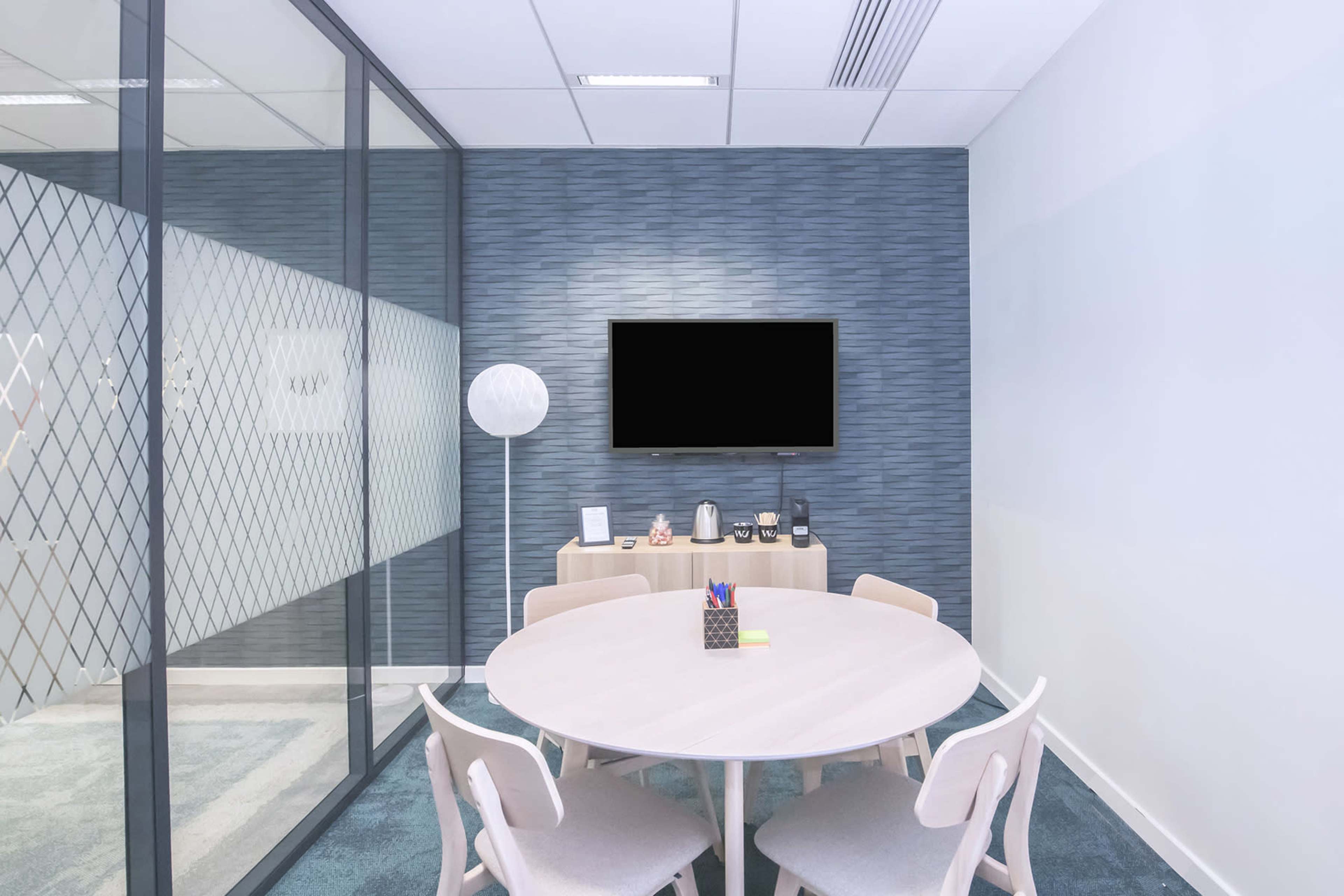A modern conference room features a round table surrounded by chairs, with a television mounted on the wall and a floor lamp in the corner.
