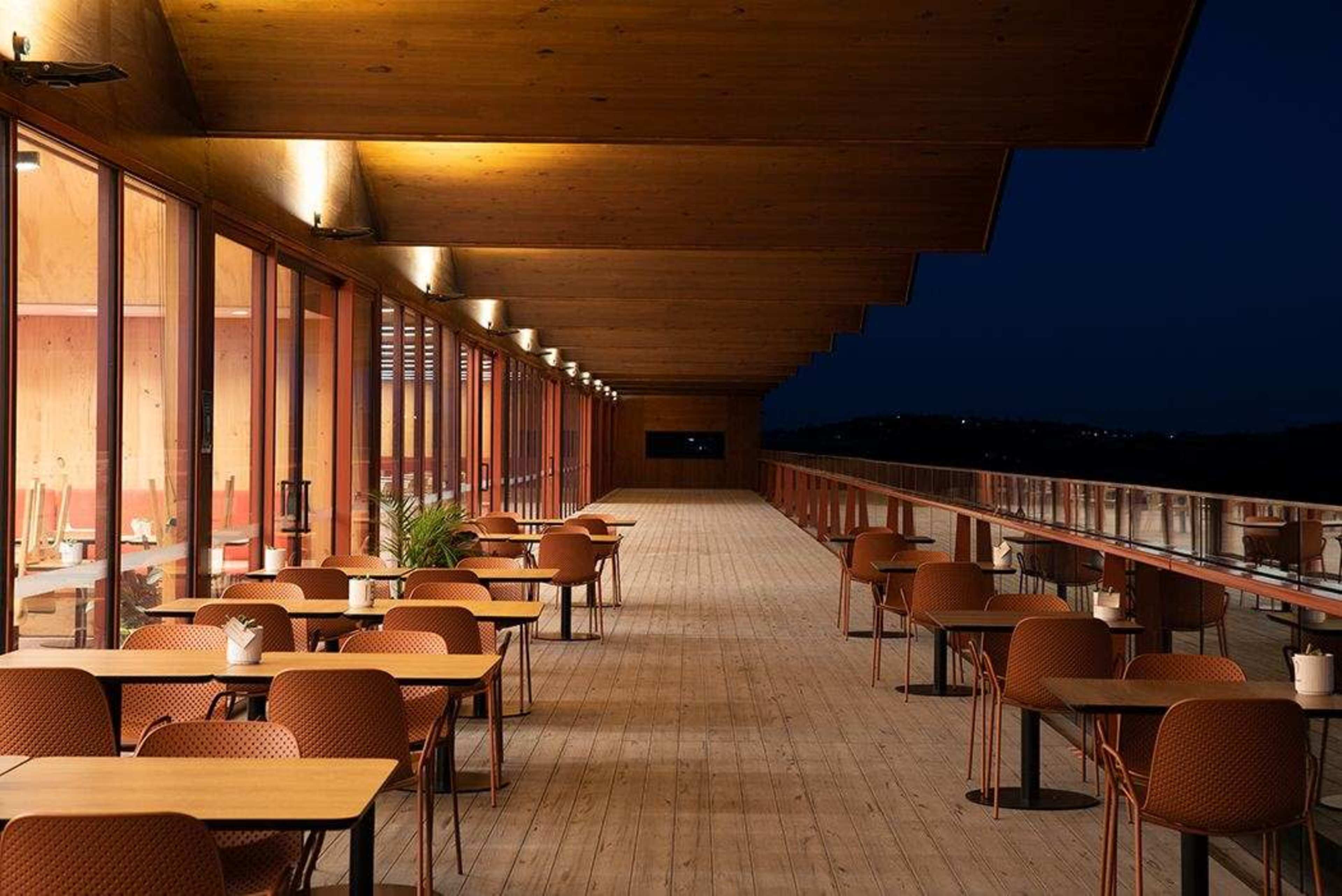 The image shows a long, illuminated veranda with wooden flooring and several tables and chairs arranged under a wooden roof at night.