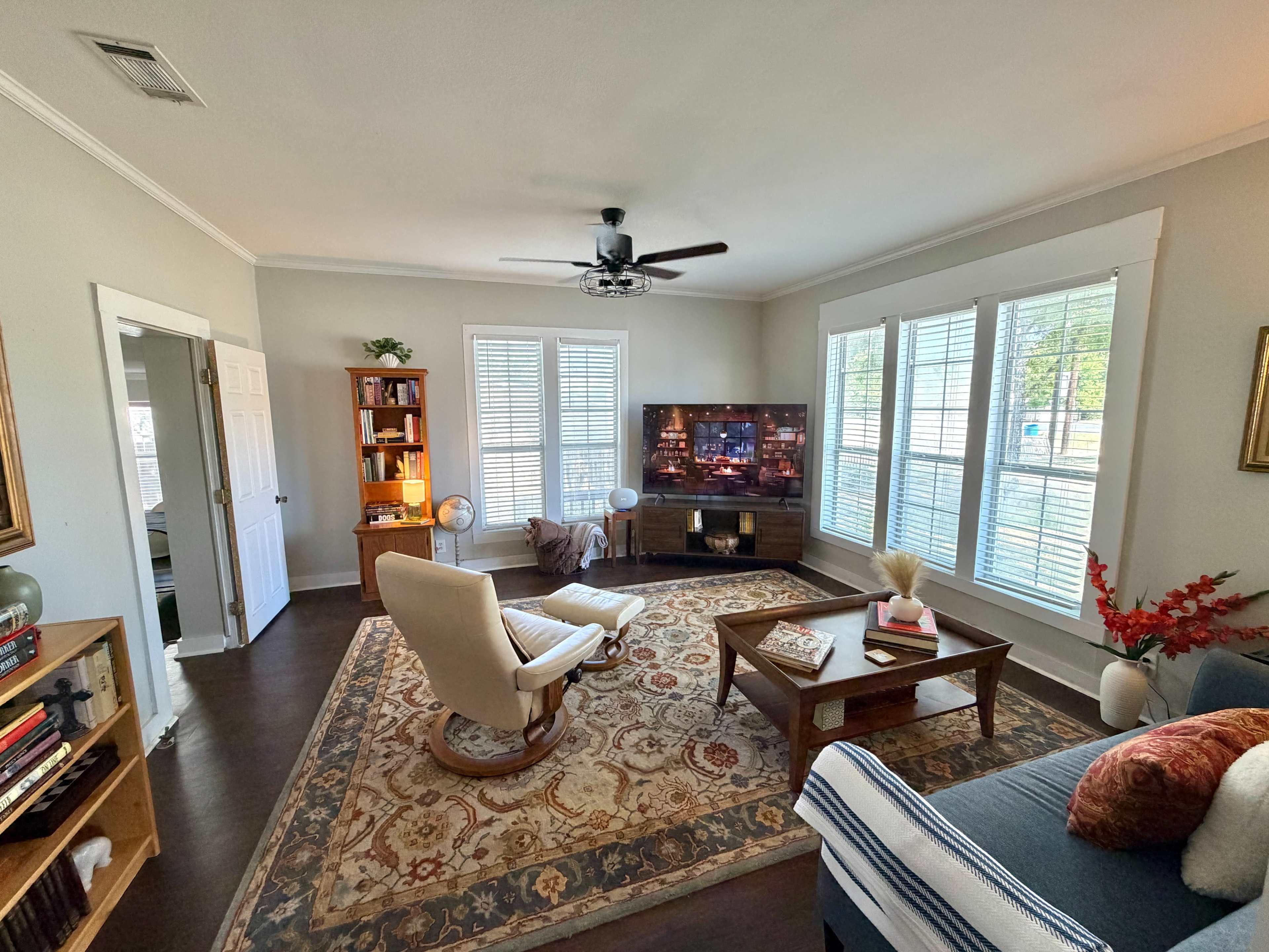 The image shows a living room with a comfortable seating area, a television on a stand, and large windows allowing natural light.