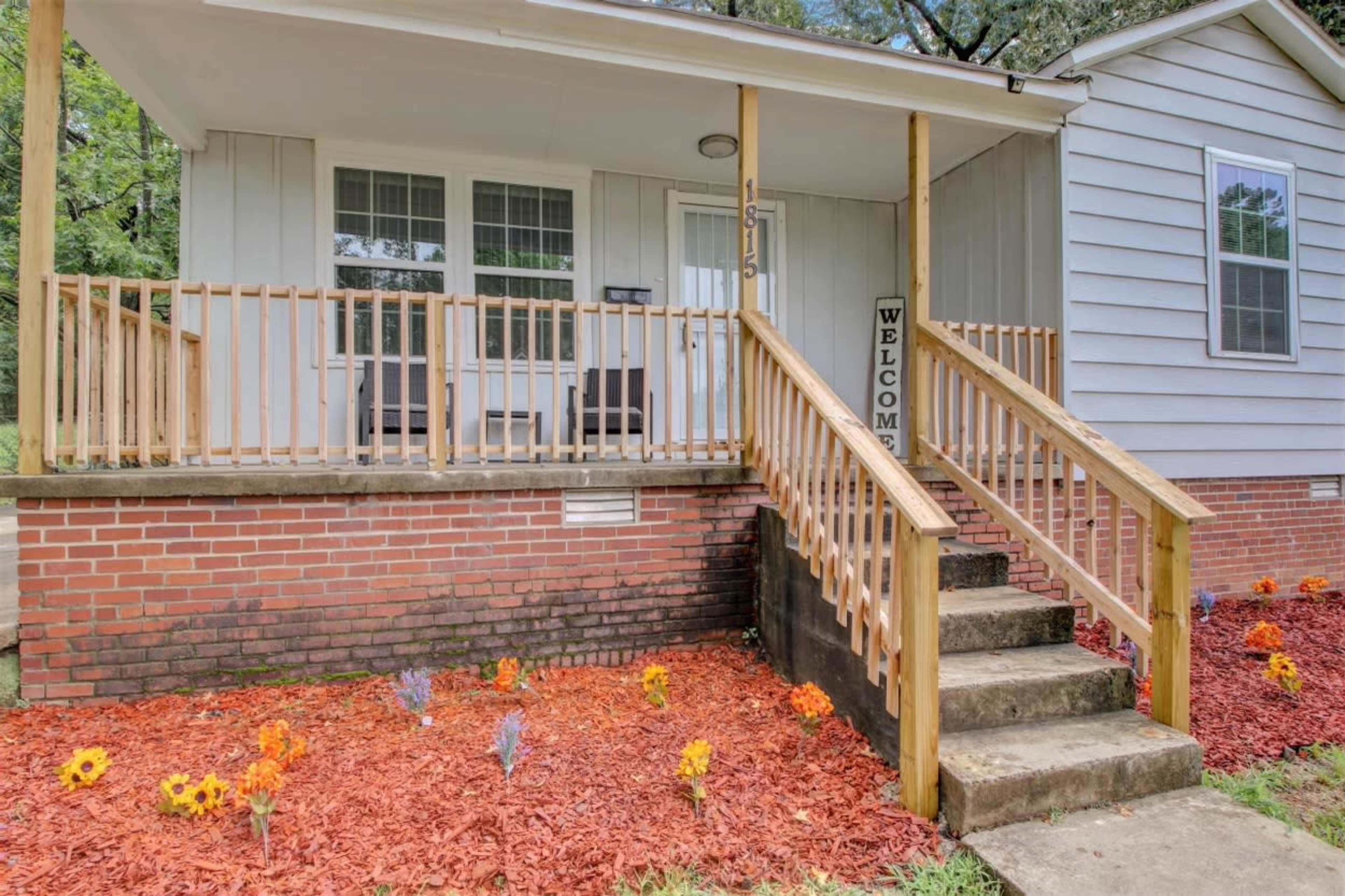 The entrance of a house with a wooden front porch, a set of stairs leading up to the door, and colorful flowers planted in the garden bed.