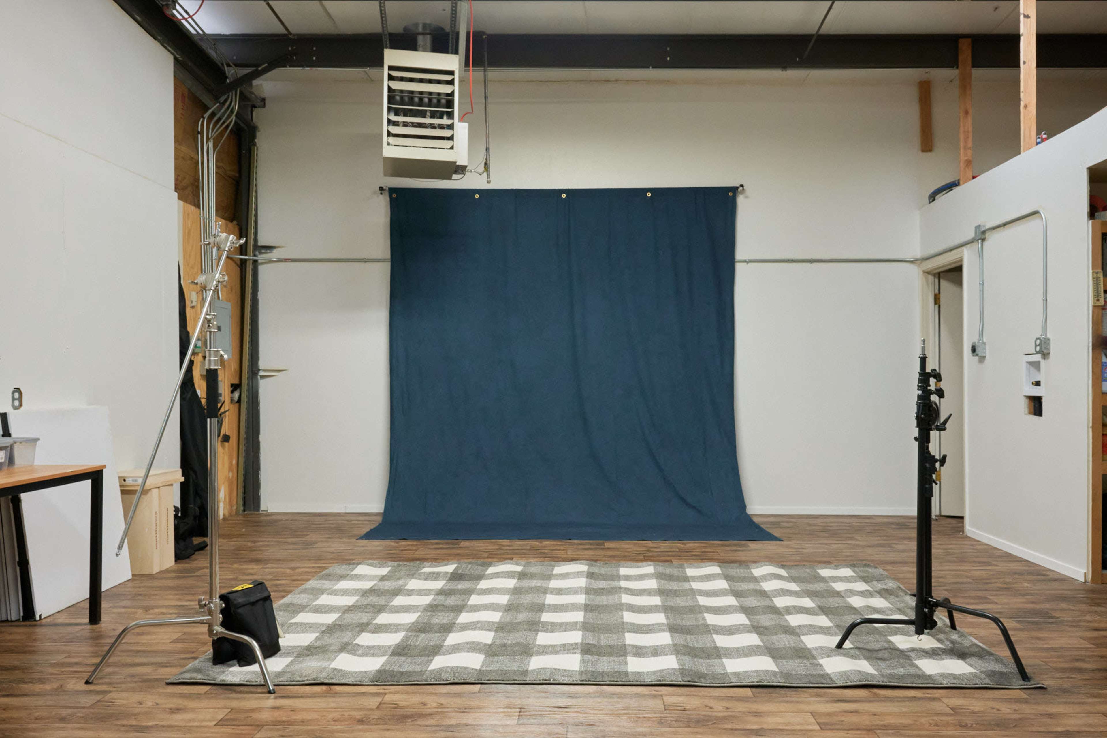 A photography studio featuring a dark backdrop, a checkered rug on the floor, and various support equipment along the walls.