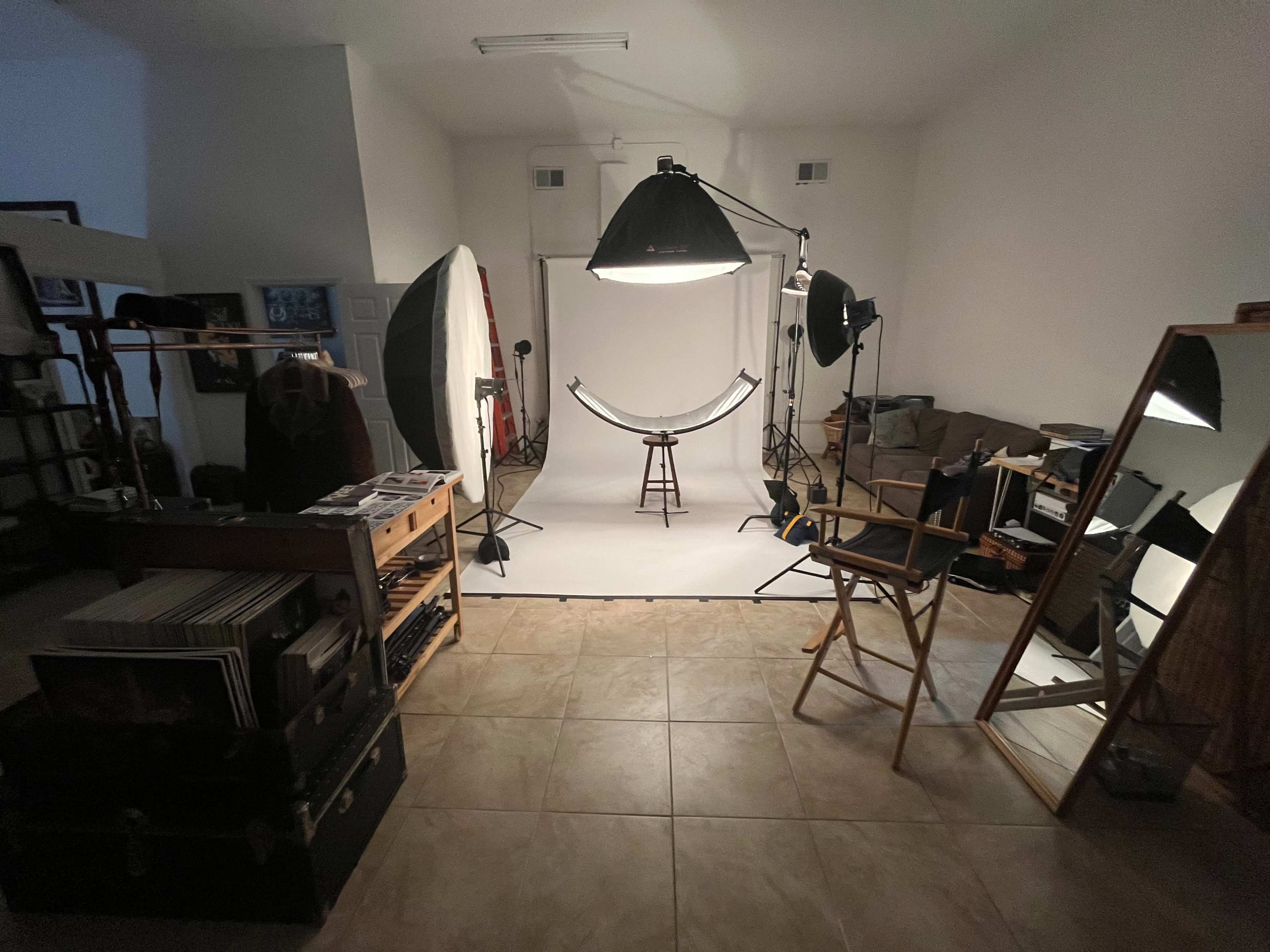 A photography studio featuring a white backdrop, various lighting equipment, a stool in the center, and a director's chair beside a large mirror.
