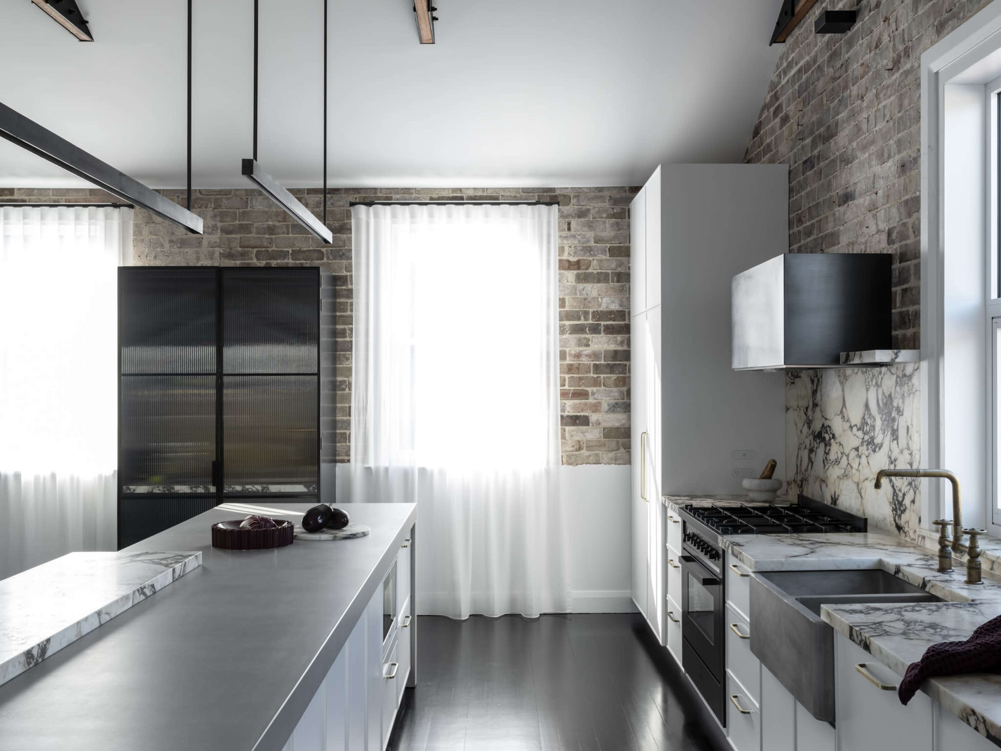 The kitchen features a blend of exposed brick walls, modern cabinetry, and a marble countertop, illuminated by natural light from a large window.
